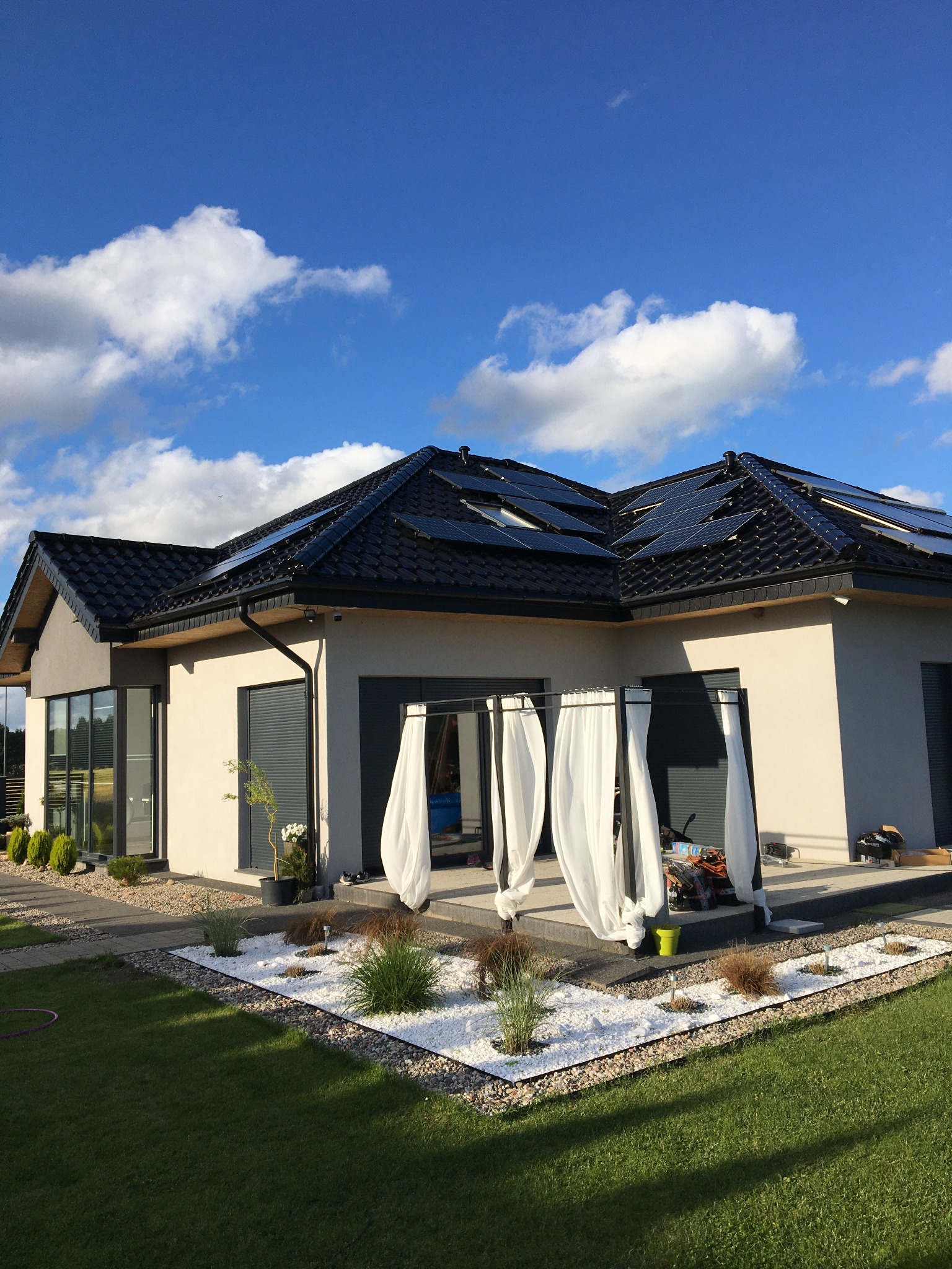 Nowoczesny dom jednorodzinny z ciemną dachówką i panelami słonecznymi, widok z ogrodu z białym kamieniem dekoracyjnym i zwiewnymi zasłonami na tarasie.