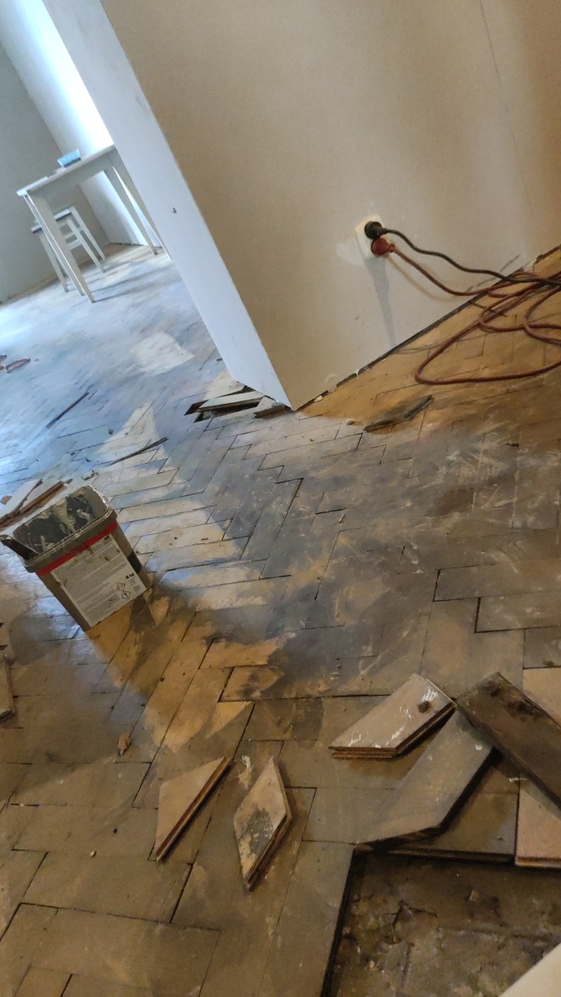 Fragment podłogi z parkietu ułożonego w jodełkę w trakcie renowacji, widoczne uszkodzenia i brakujące elementy, obok stoi opakowanie po produkcie do renowacji podłóg.