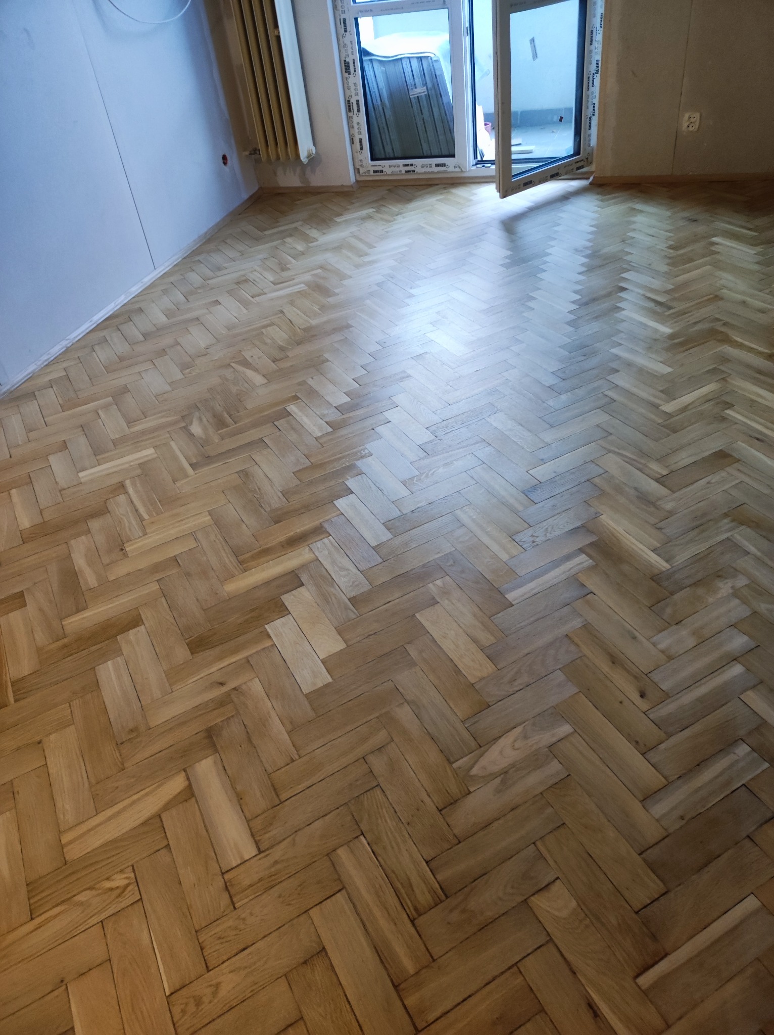 Parkiet ułożony w jodełkę po renowacji, widoczne ślady po cyklinowaniu, okno balkonowe otwarte na oścież.