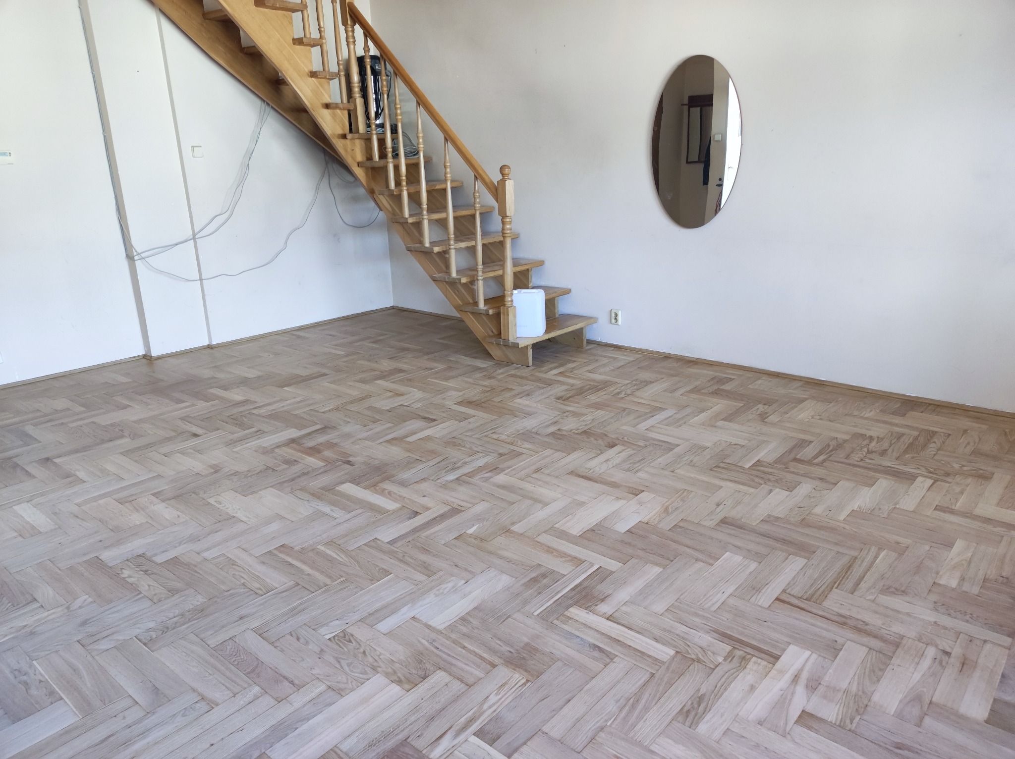 Jasna, odnowiona podłoga parkietowa ułożona w jodełkę, z drewnianymi schodami w tle, białe ściany i owalne lustro.