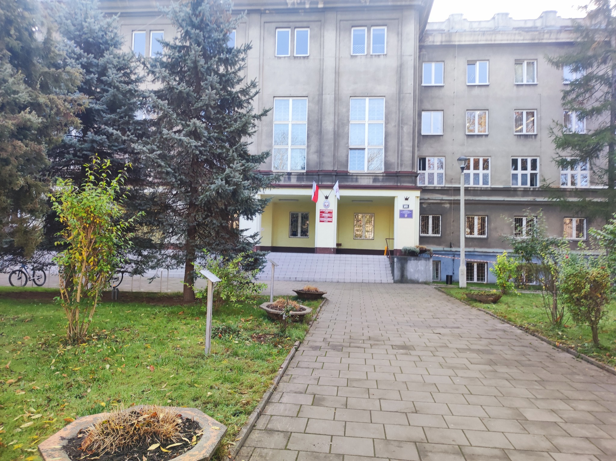 Fasada budynku użyteczności publicznej z widocznymi oknami, wejściem z flagami i otaczającą zielenią, sfotografowana w pochmurny dzień.