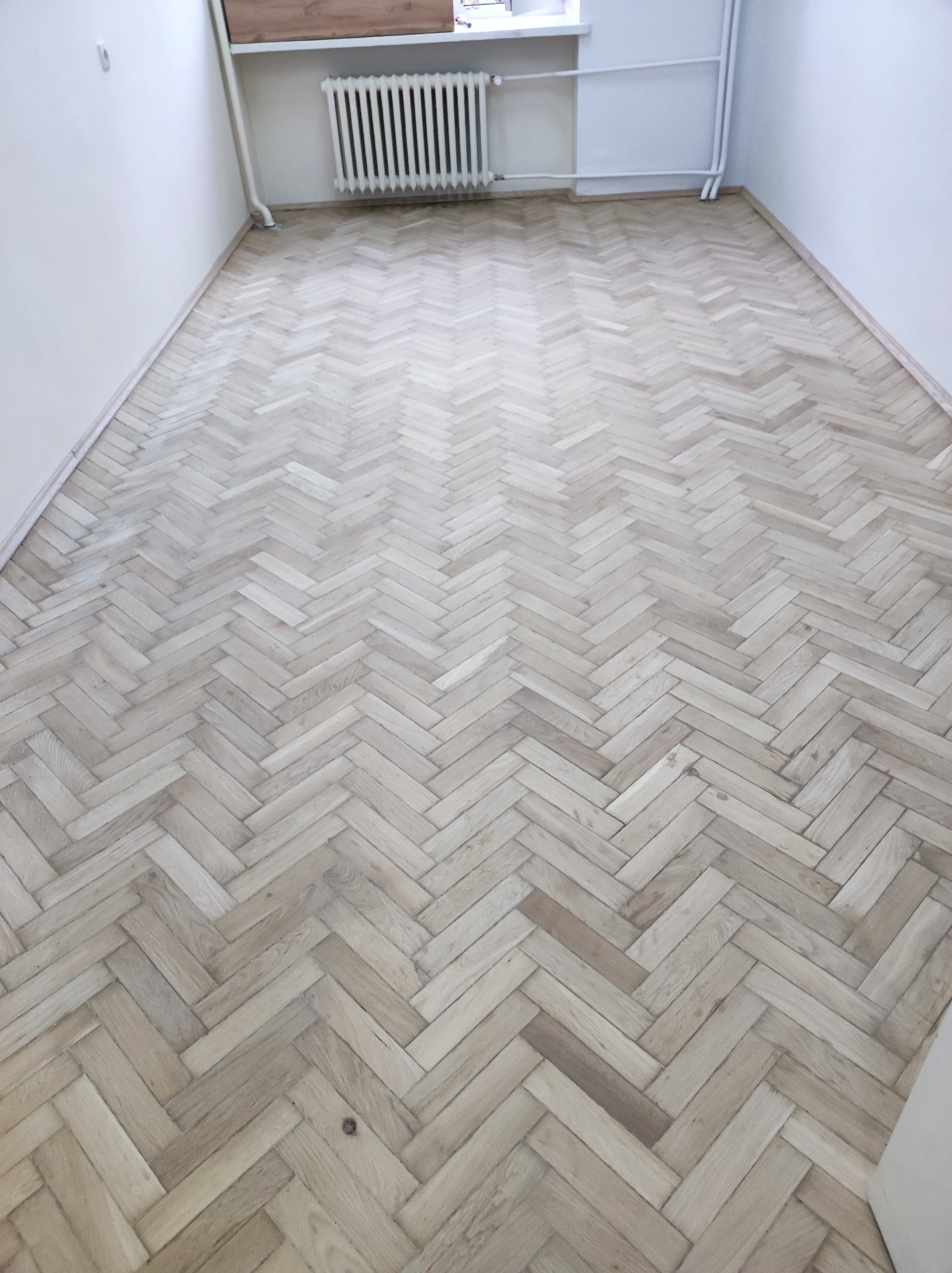 Jasna, odnowiona podłoga parkietowa ułożona w jodełkę, widok z góry, ściany pomalowane na biało, grzejnik żeliwny przy ścianie.