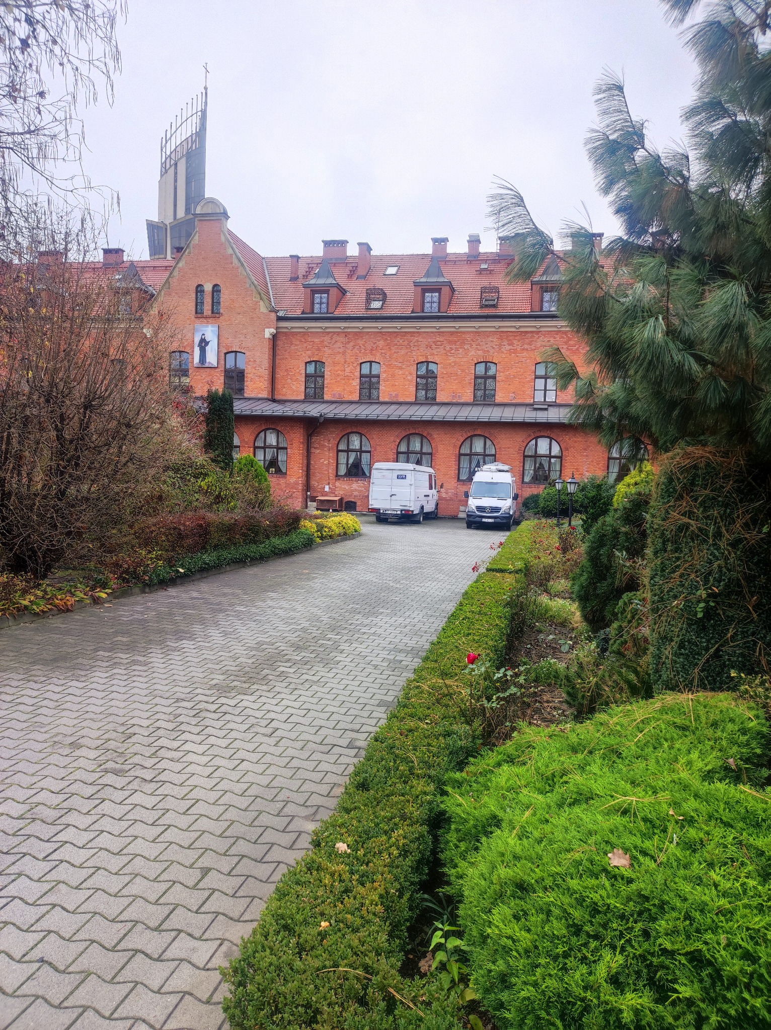 Ceglany budynek z czerwonym dachem, z przodu wybrukowany podjazd i zadbany ogród z krzewami i drzewami, dwa białe busy zaparkowane przed budynkiem.