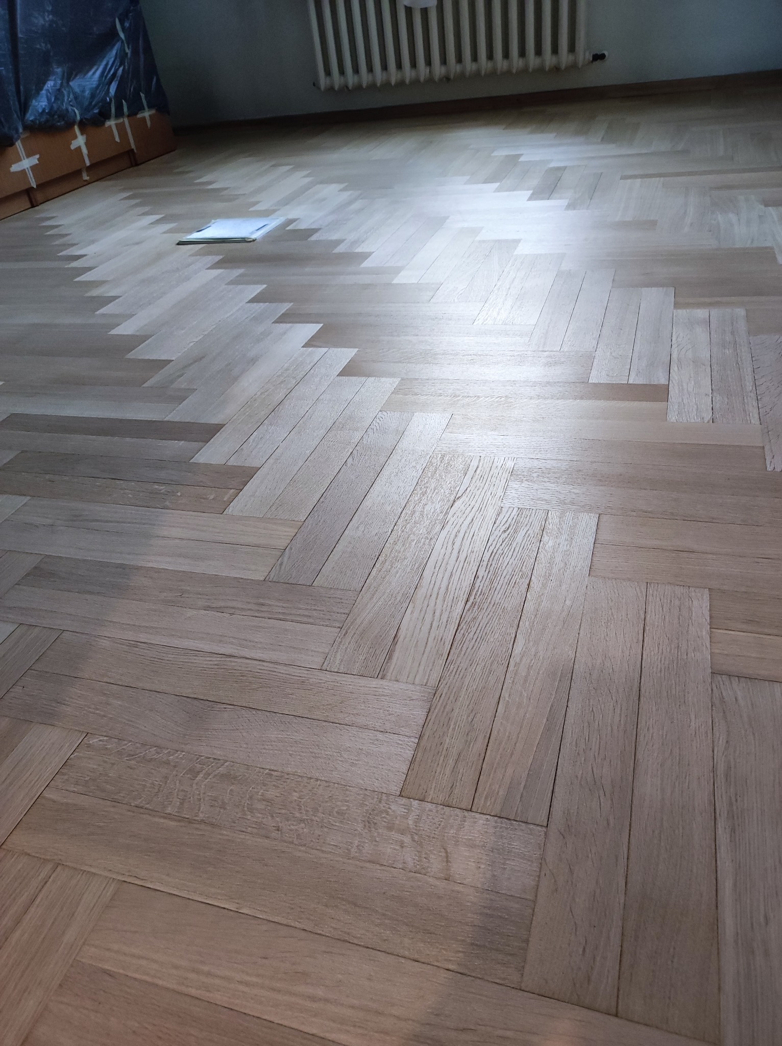 Nowo ułożony parkiet dębowy w jodełkę francuską, z widocznymi fragmentami niezabezpieczonych mebli i grzejnikiem w tle.