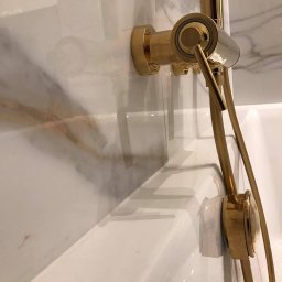 LiKo KOSTIANTYN ZHURYHORA - Złota bateria prysznicowa zamontowana na marmurowej ścianie nad białą wanną.