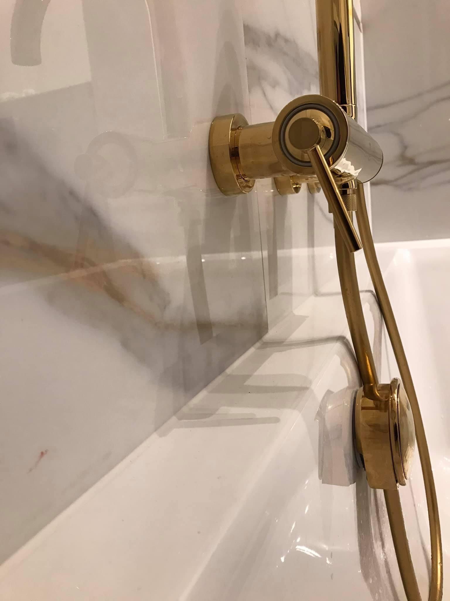 Złota bateria prysznicowa zamontowana na marmurowej ścianie nad białą wanną.