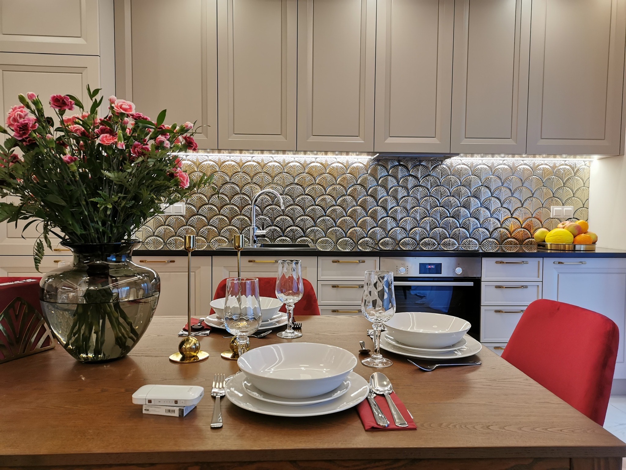 Elegancka kuchnia w odcieniach szarości z dekoracyjną mozaiką nad blatem, drewniany stół z nakryciem i bukietem różowych kwiatów w szklanym wazonie, czerwone krzesła.