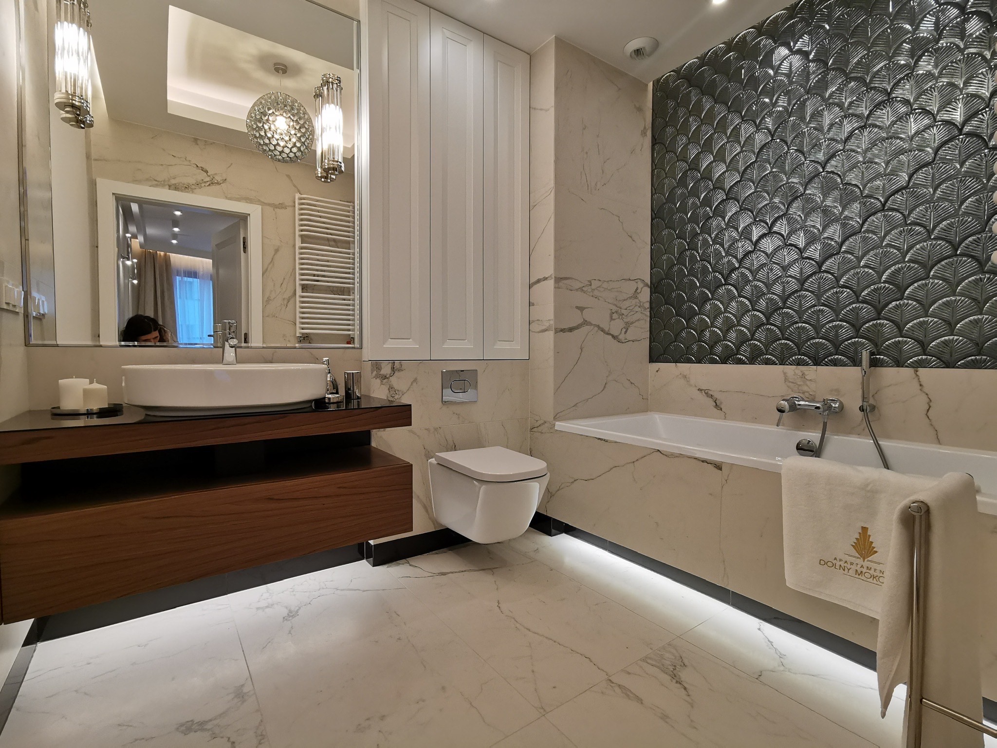 Elegancka łazienka z marmurowymi ścianami, umywalką na drewnianej szafce, toaletą podwieszaną i wanną z dekoracyjną ścianą w kształcie rybich łusek, oświetlona listwą LED przy podłodze.