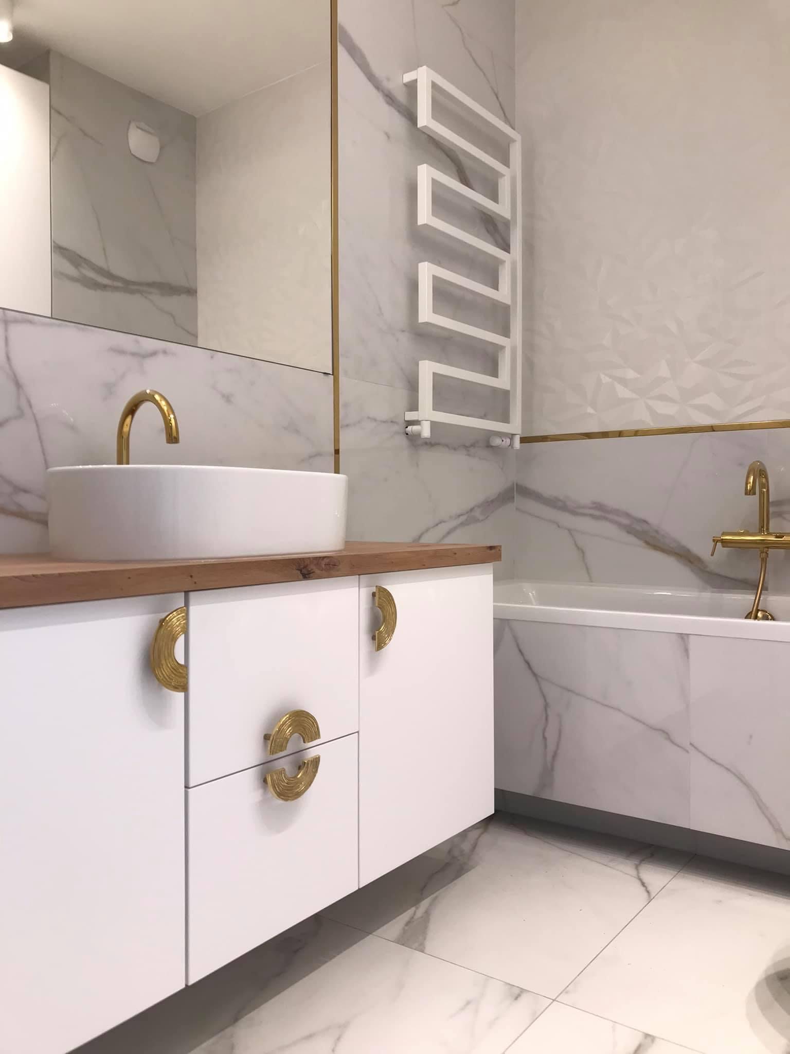 Elegancka łazienka z białymi marmurowymi płytkami, złotymi akcentami, białą umywalką nablatową i szafką z dekoracyjnymi złotymi uchwytami.