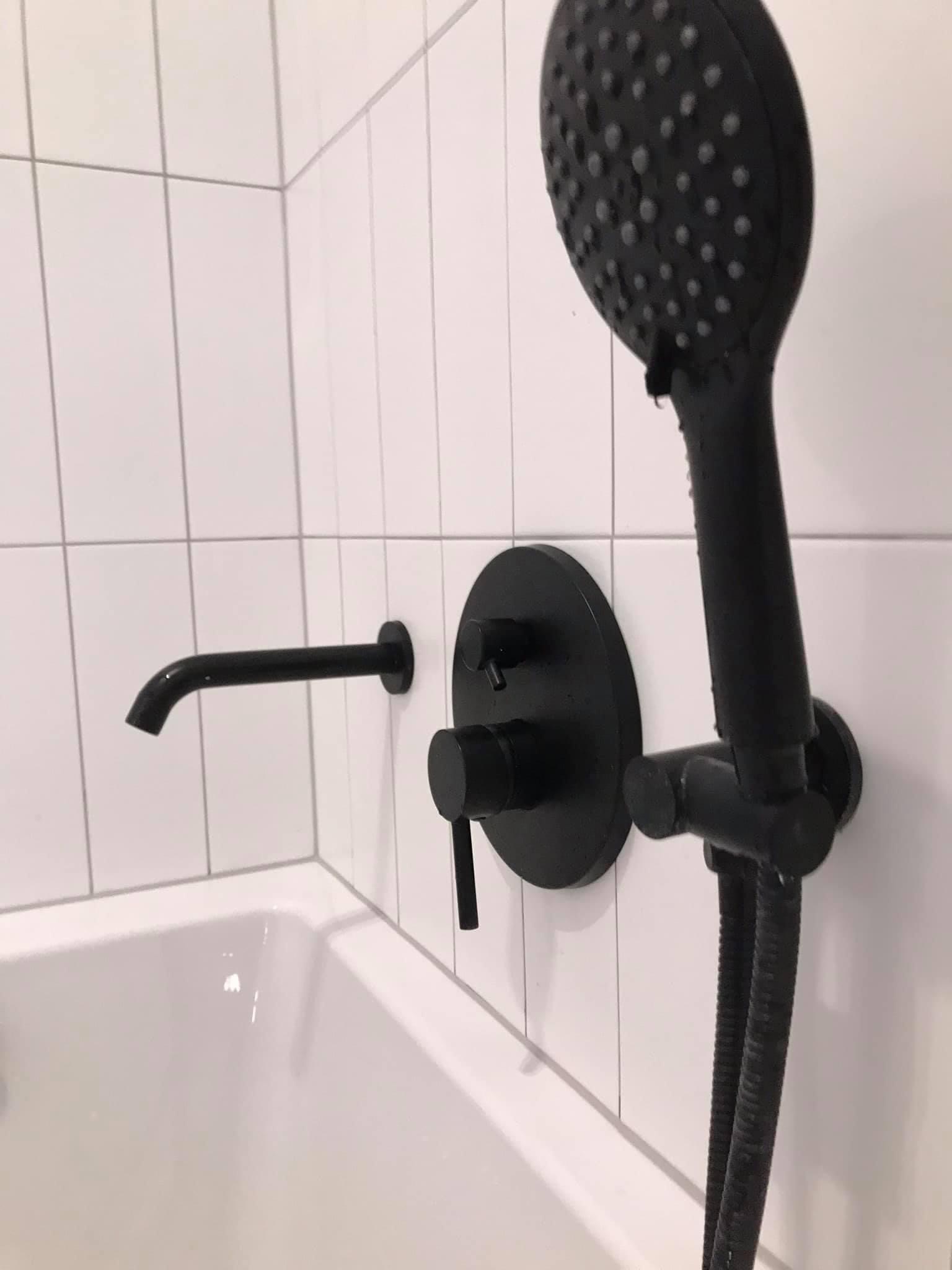 Czarna bateria wannowa natryskowa z deszczownicą, zamontowana na białych, kwadratowych płytkach, widoczna nad krawędzią wanny wypełnionej wodą.