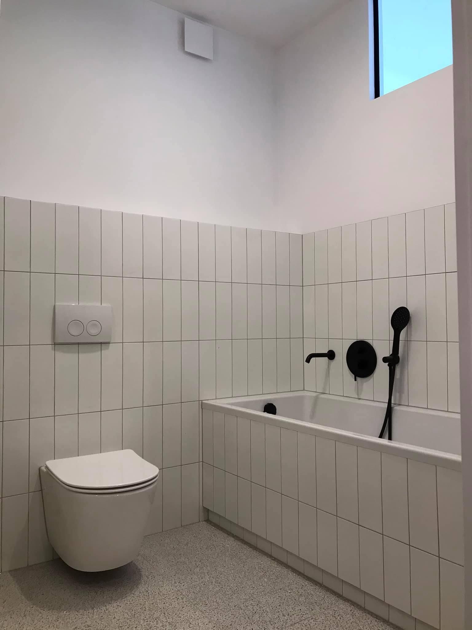 Minimalistyczna łazienka z białymi, pionowo ułożonymi płytkami, wiszącą toaletą i czarną armaturą przy wannie.