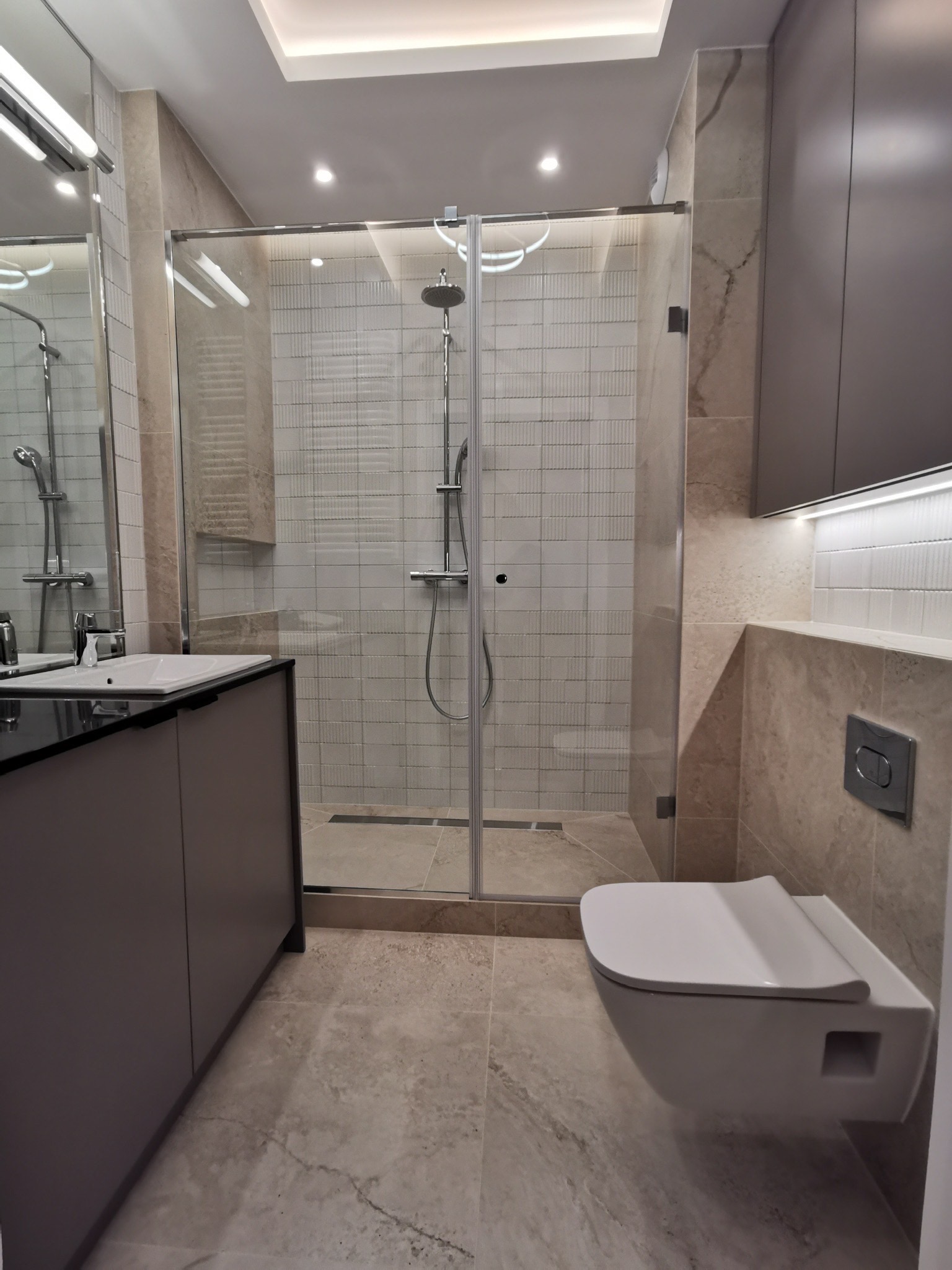 Nowoczesna łazienka z kabiną prysznicową typu walk-in, umywalką z szafką w kolorze taupe i podwieszaną toaletą, wykończona beżowymi płytkami imitującymi marmur oraz białymi płytkami strukturalnymi...