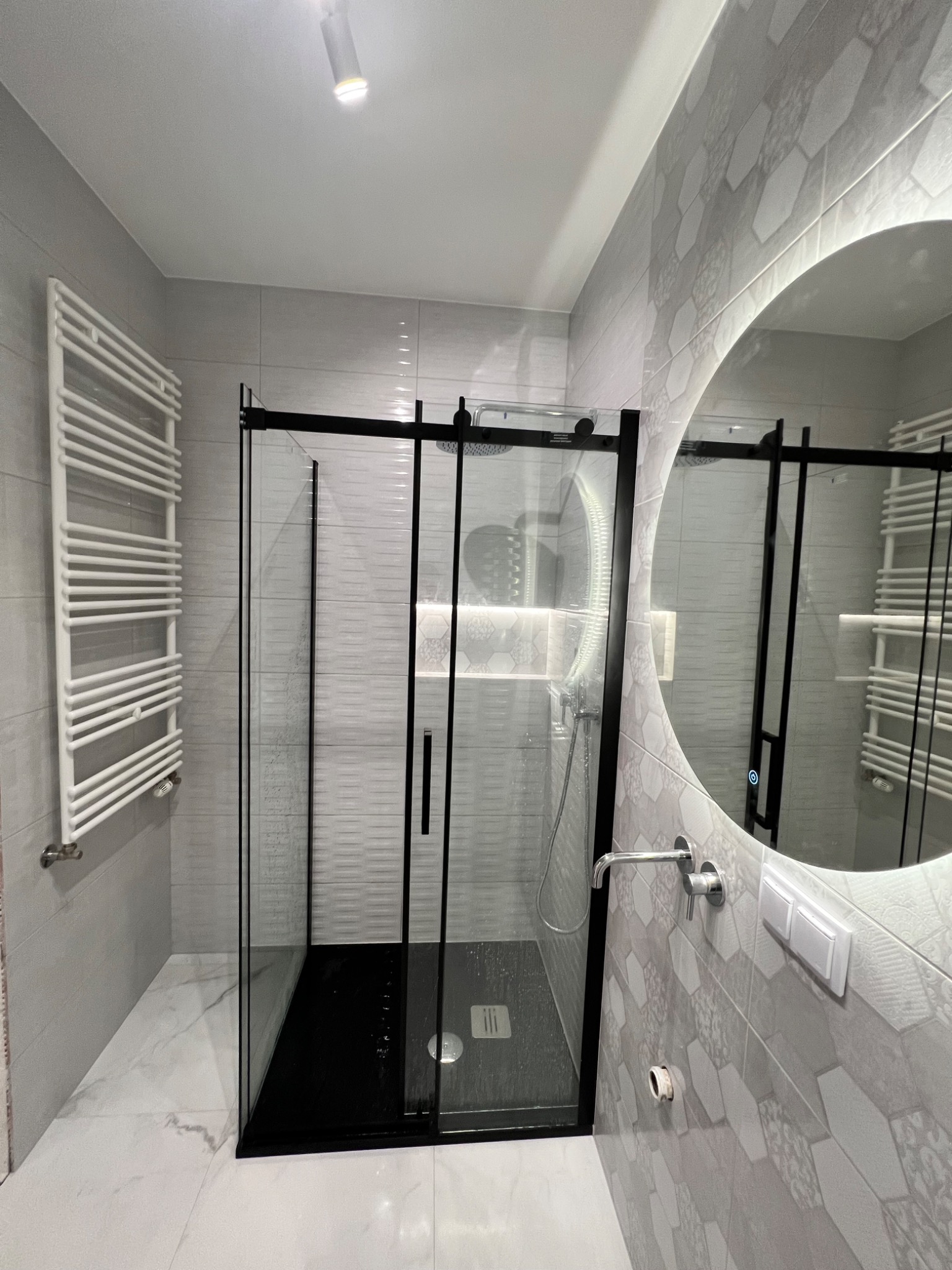 Nowoczesna łazienka z kabiną prysznicową typu walk-in z czarnymi ramami, grzejnikiem drabinkowym, okrągłym lustrem z podświetleniem LED i płytkami w geometryczny wzór.