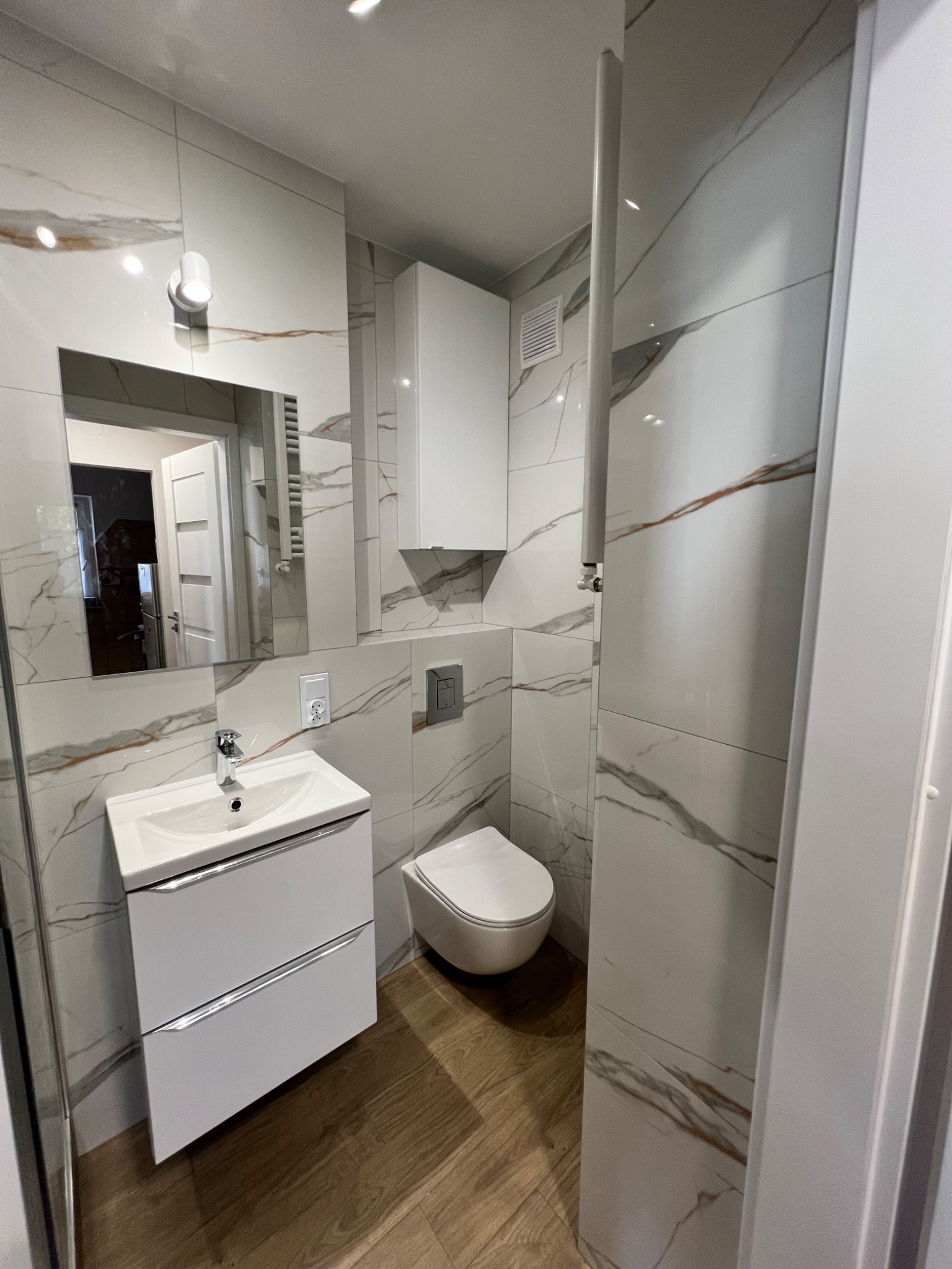 Nowoczesna, jasna łazienka z marmurowymi płytkami na ścianach, drewnopodobną podłogą, wiszącą szafką nad toaletą i umywalką z szafką, widok na lustro z odbiciem fragmentu innego pomieszczenia.