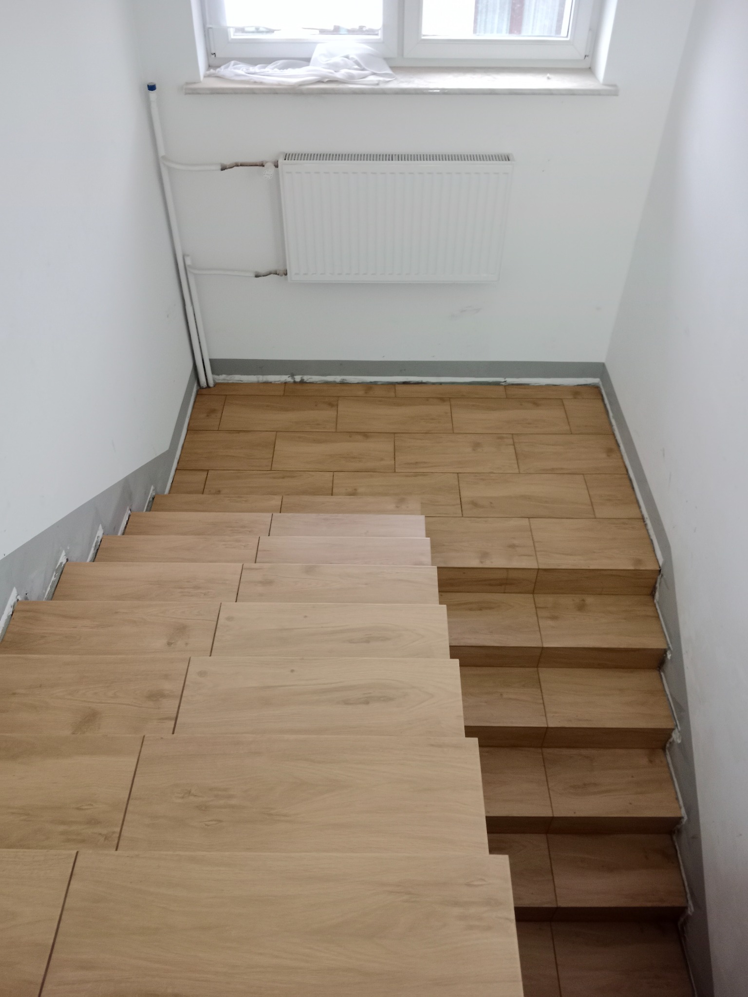 Schody wyłożone płytkami imitującymi drewno, prowadzące w dół, z widocznym grzejnikiem na ścianie i oknem u góry.