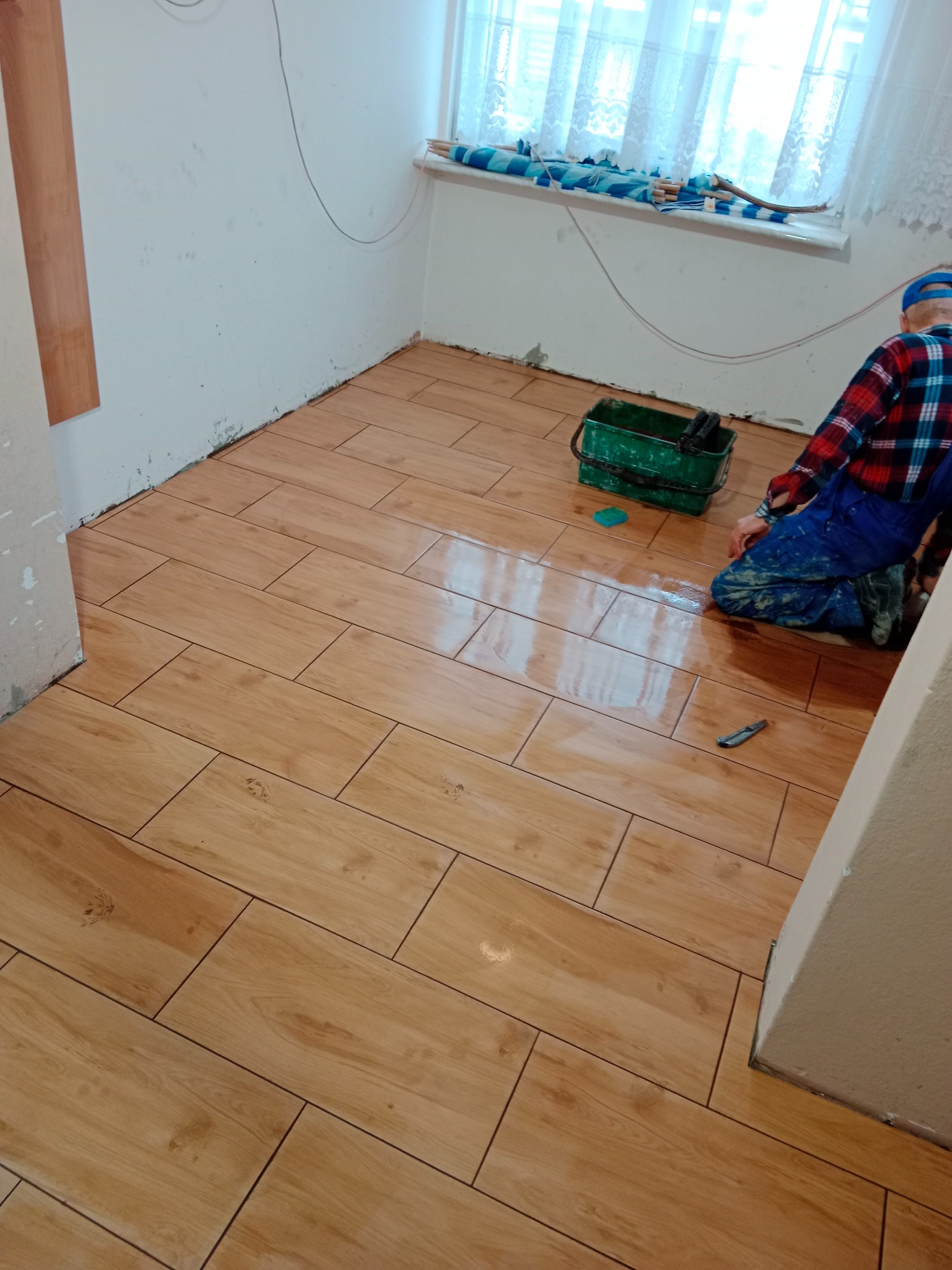 Układanie podłogi z płytek imitujących drewno w małym pokoju; widoczny pracownik w ubraniu roboczym, zielone wiadro z narzędziami i nóż do cięcia płytek.
