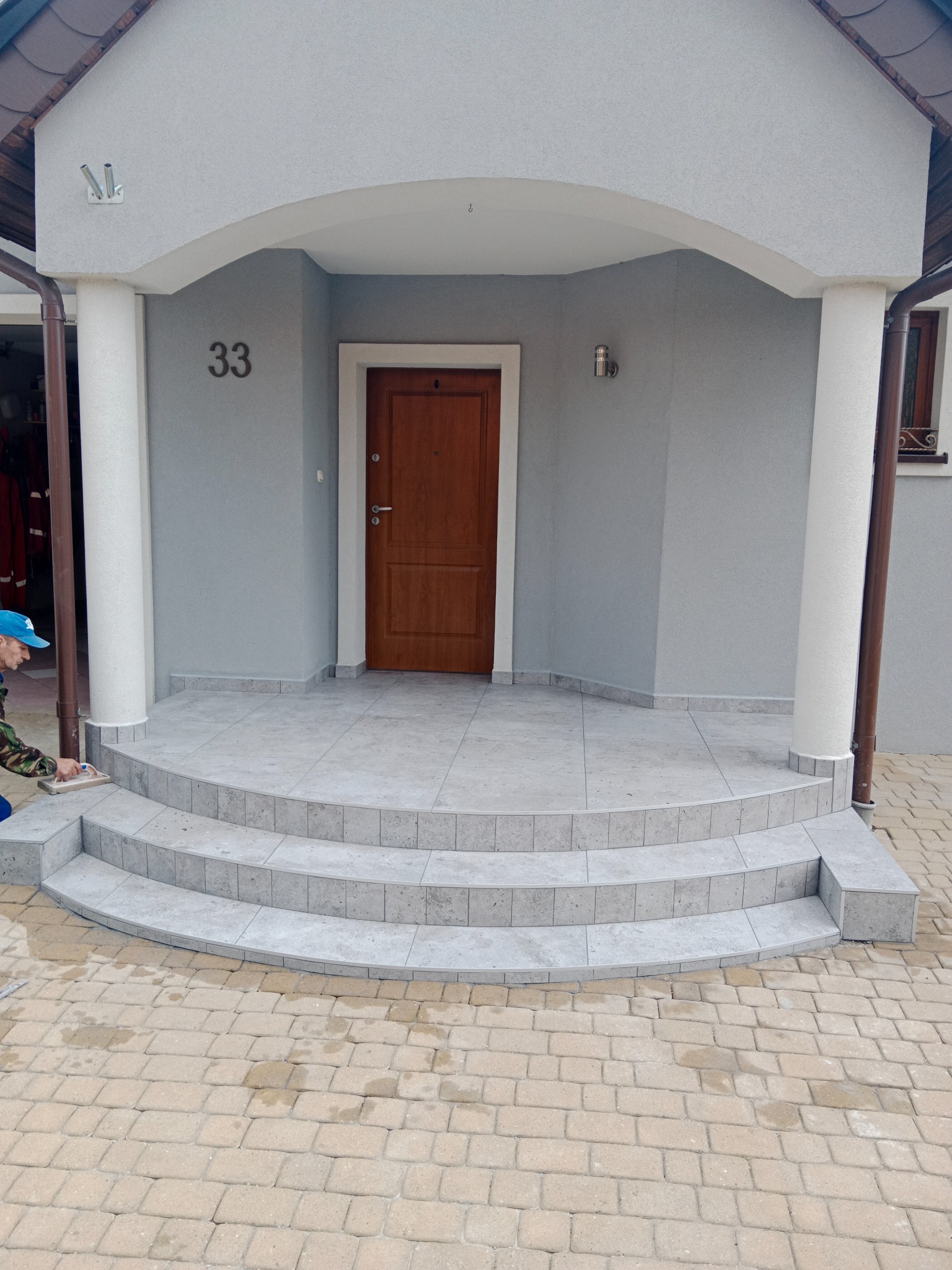Wykończenie schodów wejściowych do domu z szarych płytek, widoczny pracownik zacierający fugi na ostatnim stopniu, numer domu 33.