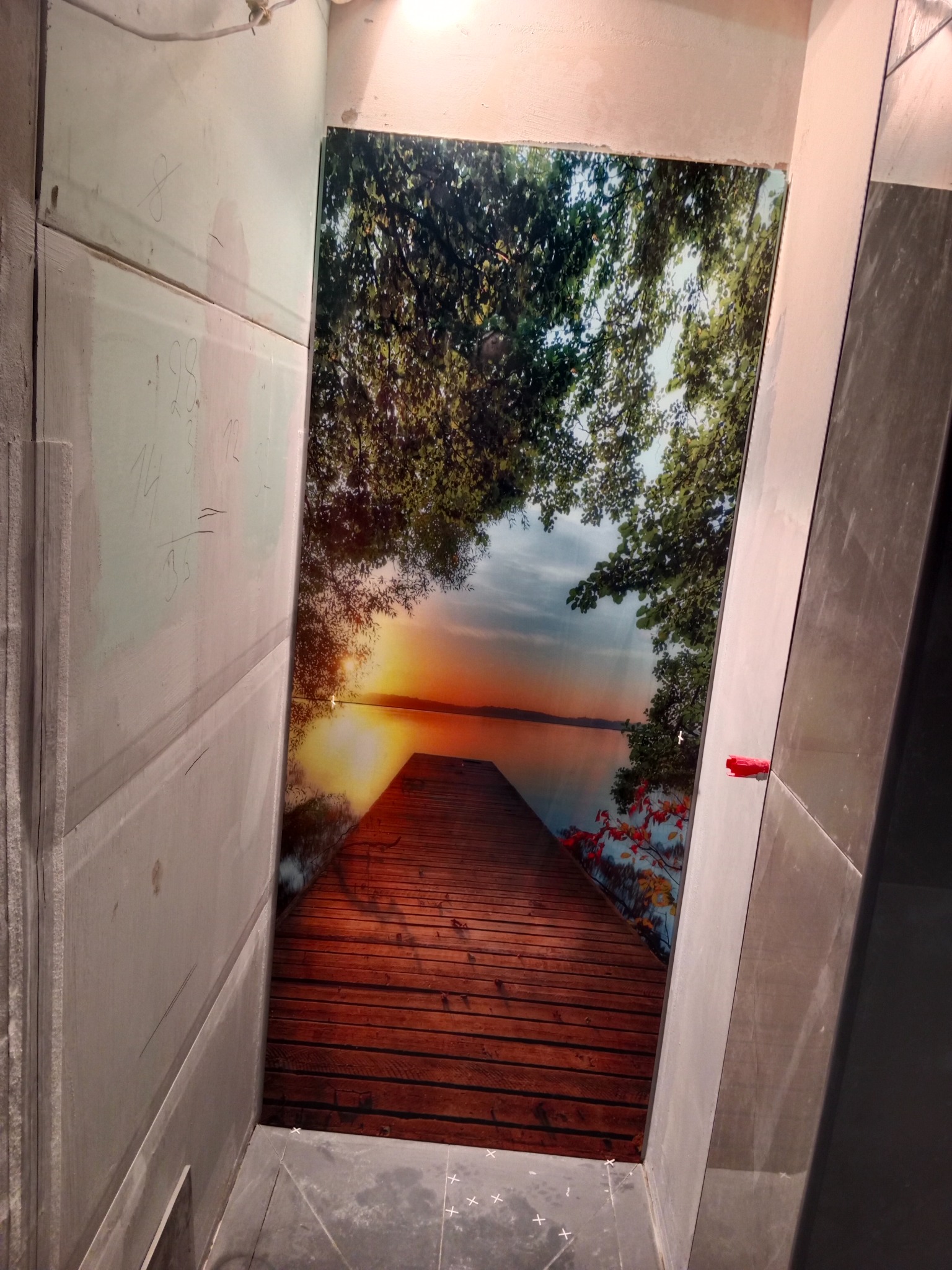 Wąska łazienka z fototapetą przedstawiającą drewniany pomost nad jeziorem o zachodzie słońca, widoczne surowe ściany przygotowane do wykończenia.
