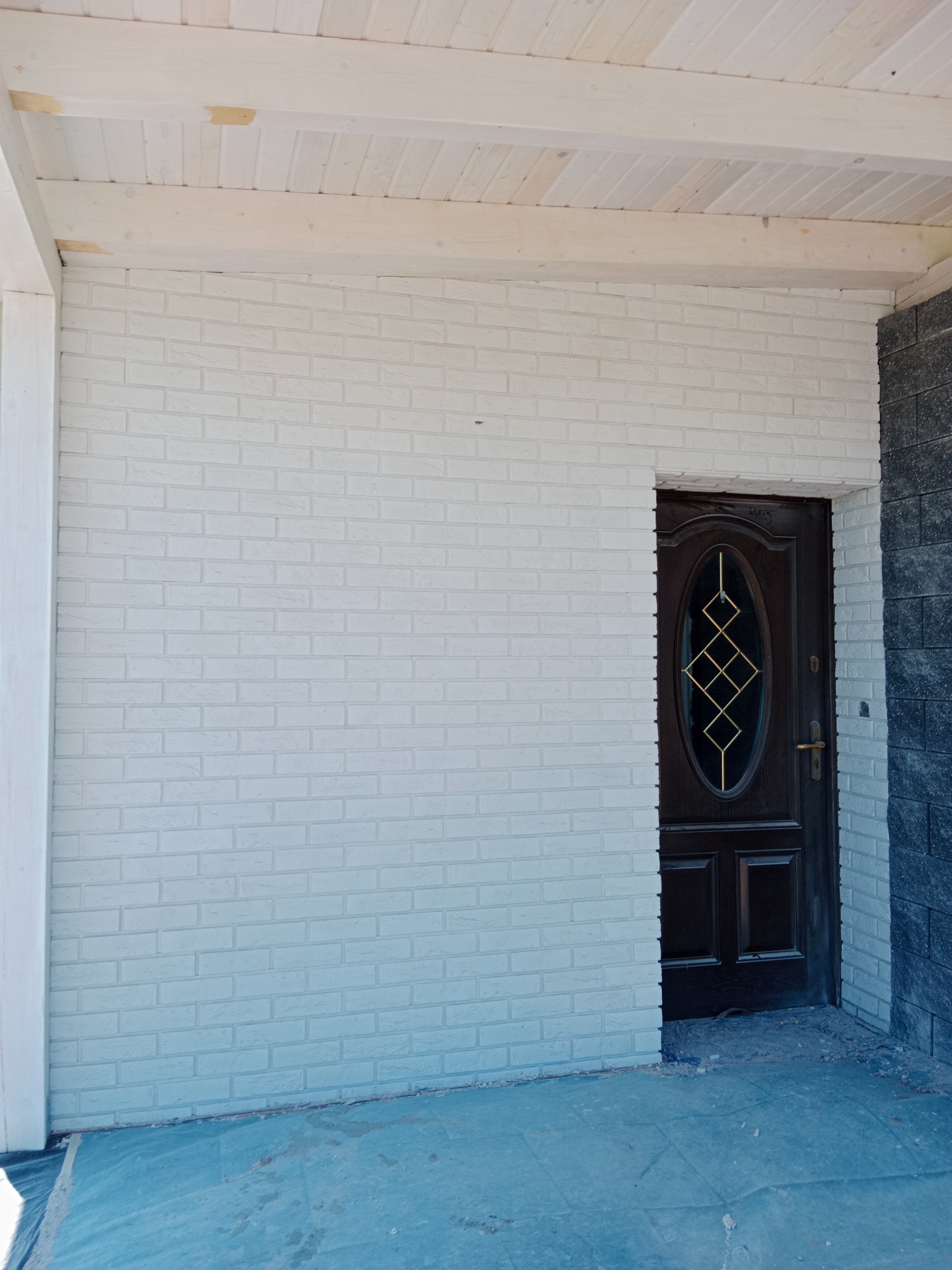 Biała ściana z cegły klinkierowej na zewnątrz budynku, z ciemnymi drzwiami wejściowymi z owalnym przeszkleniem i elementami dekoracyjnymi, oraz fragmentem ściany wykończonej ciemnoszarym kamieniem...