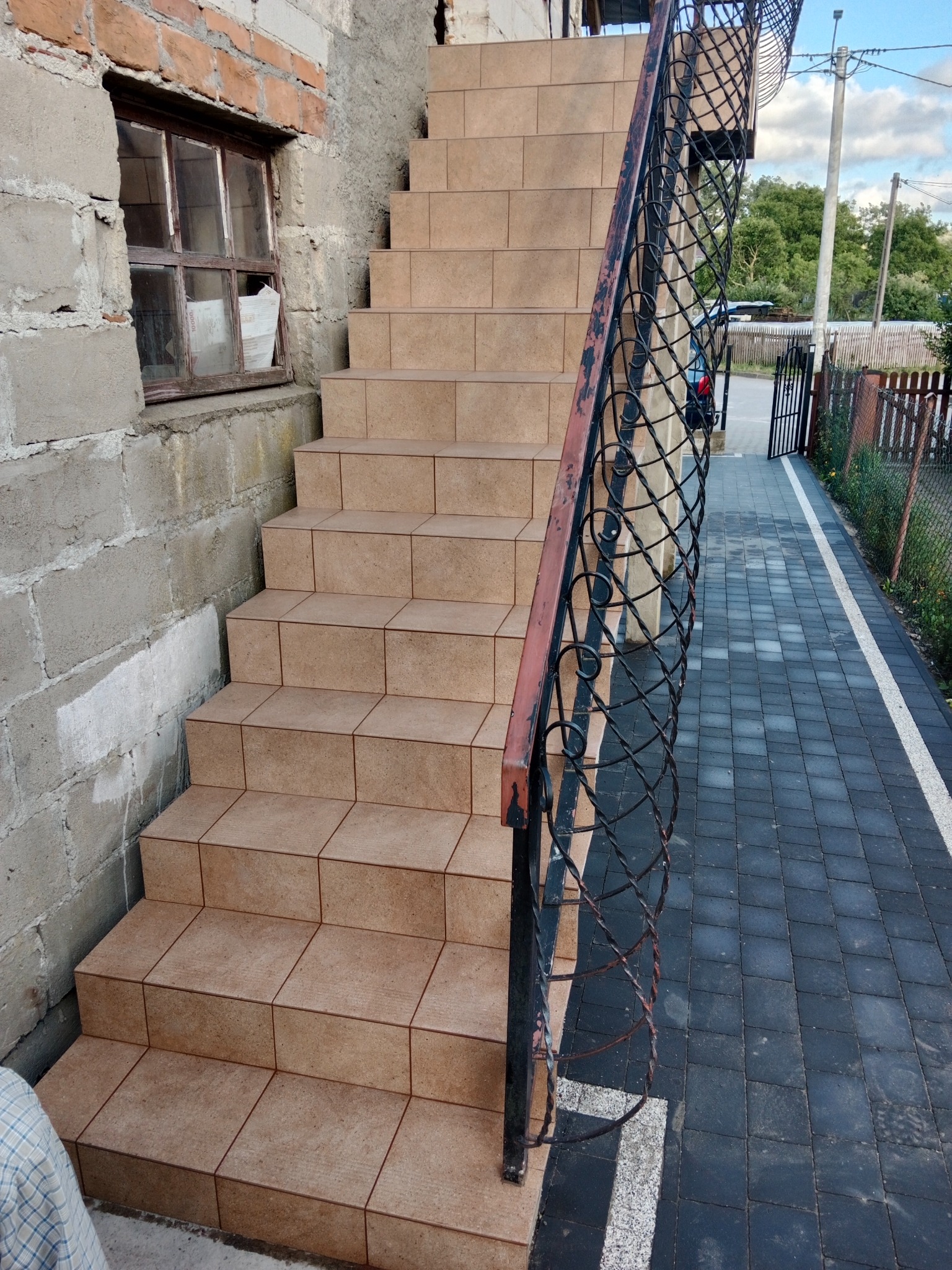 Zewnętrzne schody wyłożone beżowymi płytkami, z metalową, ozdobną balustradą, prowadzące do wejścia. Widoczna elewacja budynku z oknem i kostka brukowa na podwórku.