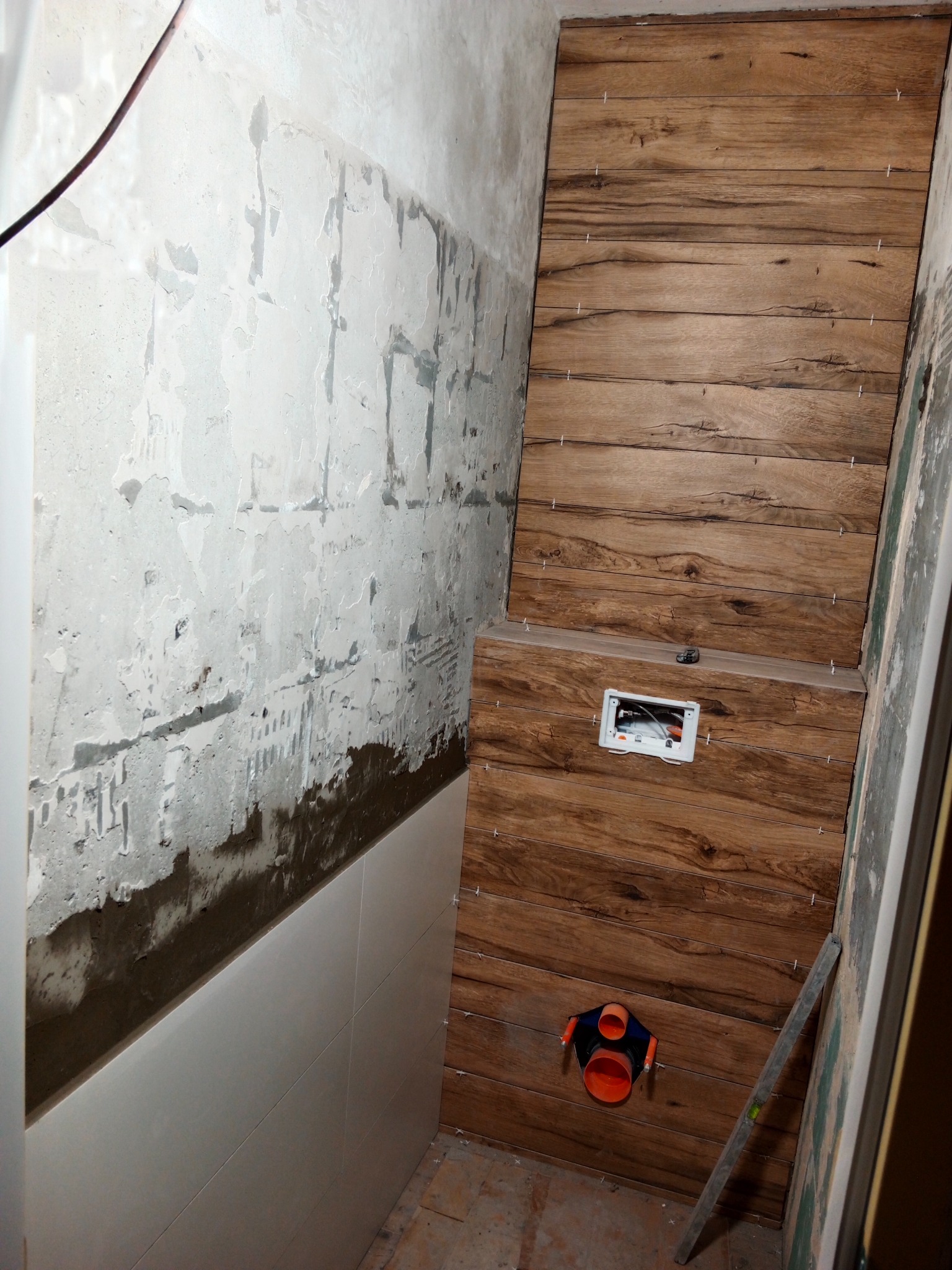 Montaż drewnopodobnych płytek na ścianie w łazience, widoczny fragment instalacji sanitarnej oraz poziomica oparta o ścianę, surowe ściany w tle.