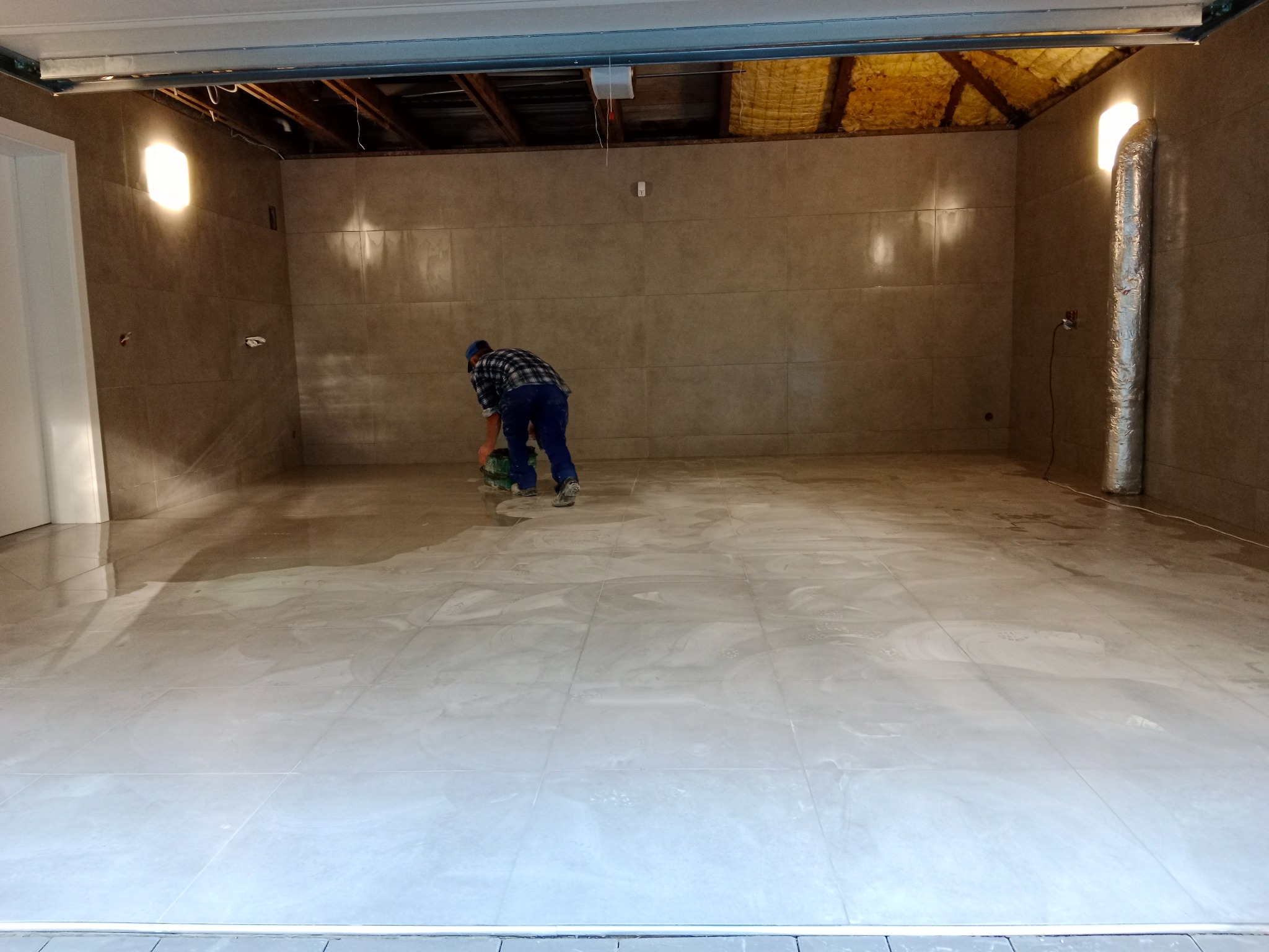 Układanie jasnych, marmurkowych płytek podłogowych w pomieszczeniu z szarymi ścianami; pracownik w niebieskim kombinezonie i czapce, szlifuje fugi przy pomocy szlifierki.