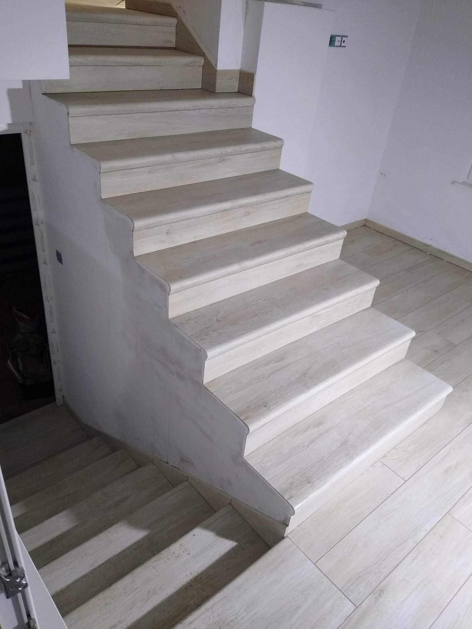 Wykończone schody wewnętrzne z płytkami imitującymi drewno, z zaokrąglonymi krawędziami, dopasowane do podłogi, widoczne niedoskonałości wykończenia ściany przy schodach.