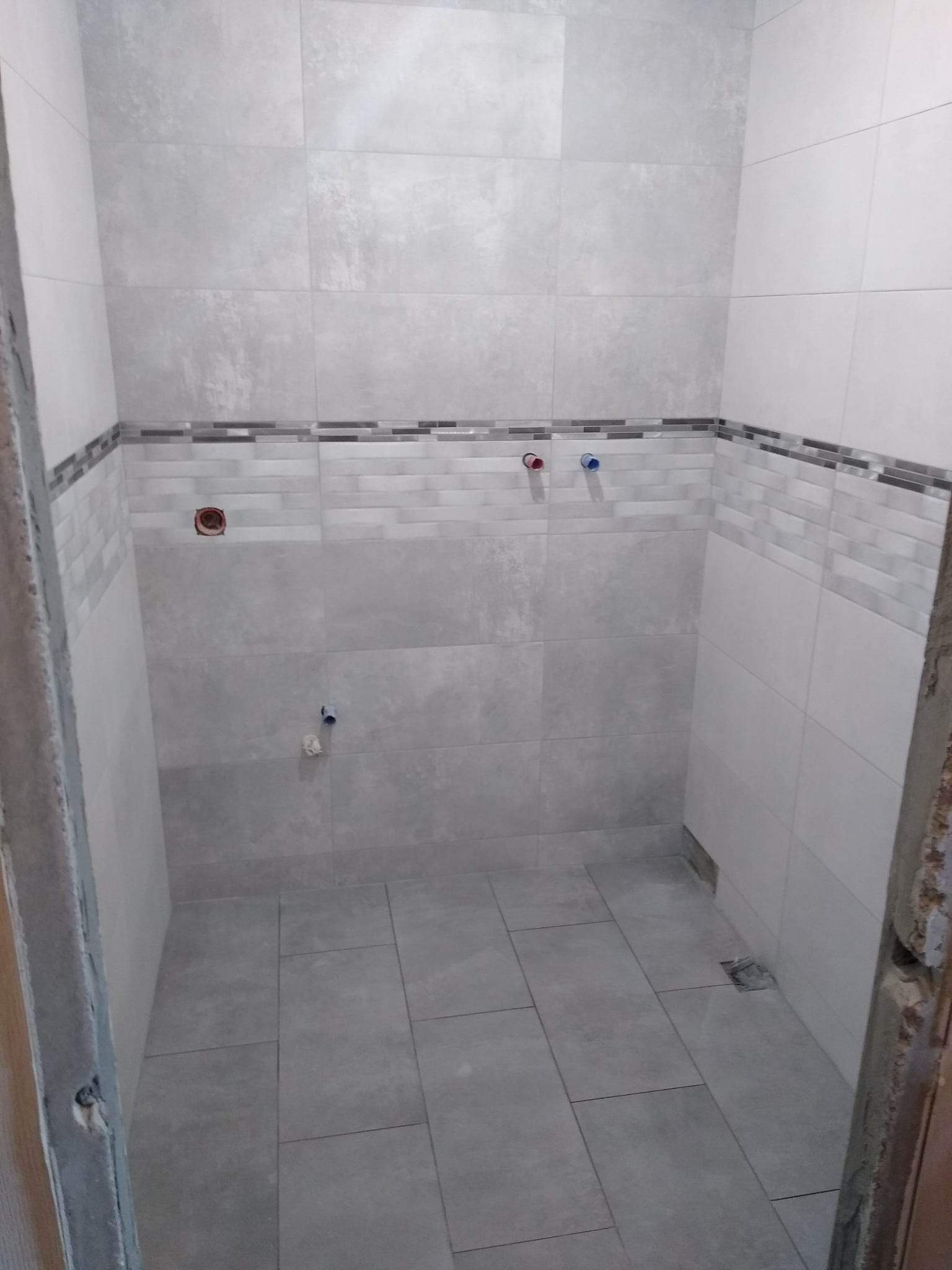 Wykończona łazienka z szarymi płytkami imitującymi beton, widoczne podejścia wodne i dekoracyjny pas płytek mozaikowych pod sufitem, ujęcie z poziomu podłogi.