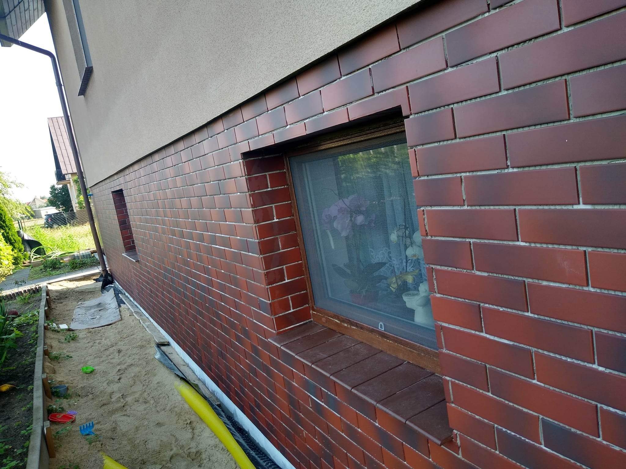 Elewacja domu wykończona czerwoną cegłą klinkierową wokół okien; widoczny fragment ogrodu z piaskiem i żółtymi rurami.