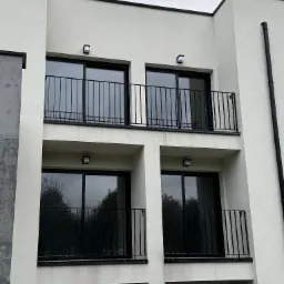 Fasada nowoczesnego, trzypiętrowego budynku z balkonami zabezpieczonymi czarnymi, metalowymi barierkami, oświetlonymi przez kwadratowe lampy ścienne.