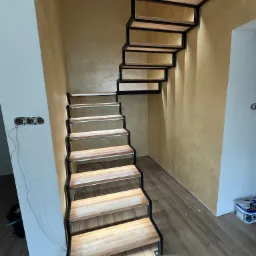 Nowoczesne schody z drewnianymi stopniami i czarną metalową konstrukcją w jasnym wnętrzu. Widoczne gniazdka elektryczne i faktura ściany.