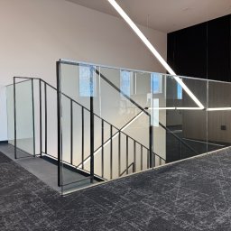 STAL KRAFT - Nowoczesne schody z balustradą szklaną i czarnymi poręczami, widok z góry na szary dywan. Minimalistyczny design, oświetlenie LED. Widoczne odbicie drzew w szkle.