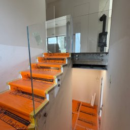 STAL KRAFT - Szklana balustrada na schodach w trakcie montażu, zabezpieczone pomarańczową folią ochronną. Widoczne elementy mocujące i fragment wnętrza z kominkiem.