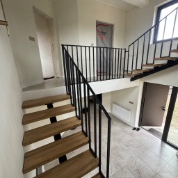 STAL KRAFT - Nowoczesne schody z drewnianymi stopniami i czarną metalową balustradą w jasnym wnętrzu. Widok z góry na schody i wejście do domu. Minimalistyczny design.