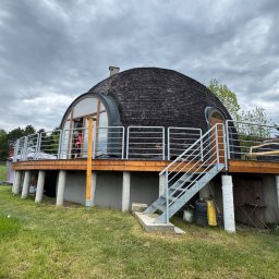 STAL KRAFT - Nietypowy dom w kształcie kopuły z drewnianym dachem i metalową balustradą, otoczony zielonym trawnikiem pod pochmurnym niebem. Schody prowadzą na drewniany taras.