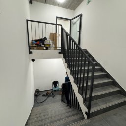 STAL KRAFT - Klatka schodowa w nowoczesnym budynku z szarymi schodami i czarną, metalową balustradą. Widoczne narzędzia i materiały budowlane na podeście.