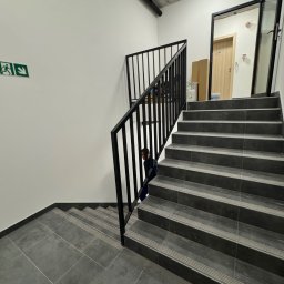 STAL KRAFT - Nowoczesne schody z szarych płytek z czarną, prostą balustradą metalową w jasnym wnętrzu budynku. Widoczny fragment osoby na schodach i znak ewakuacyjny na ścianie.