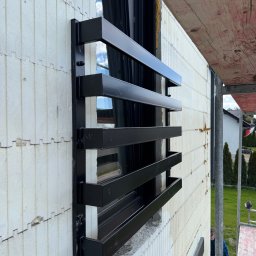 STAL KRAFT - Czarna, metalowa krata okienna, zamontowana na białej ścianie budynku w trakcie budowy, widoczna z boku, z fragmentem rusztowania w prawym górnym rogu.