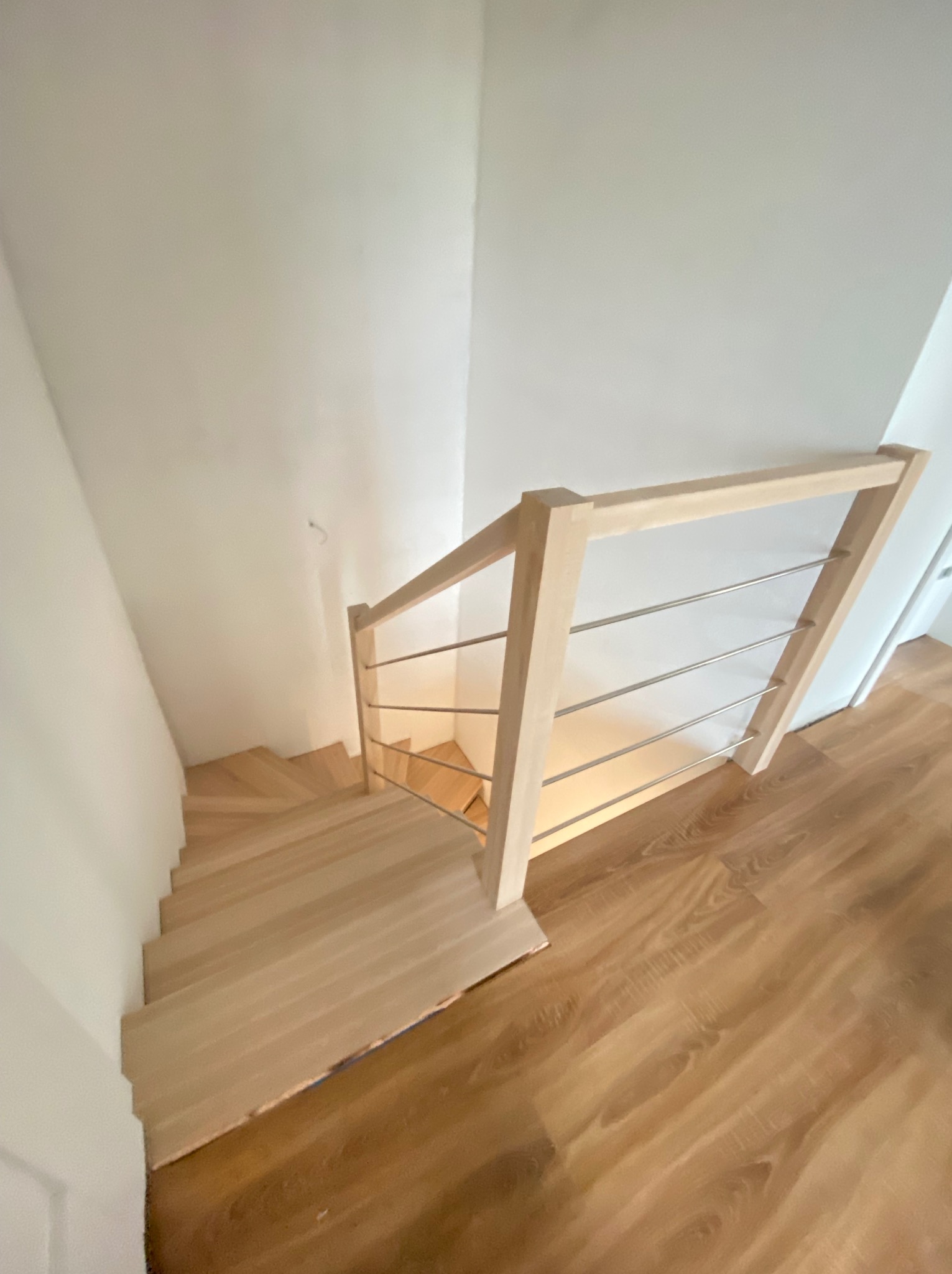 Widok z góry na jasne, drewniane schody z prostą, drewnianą balustradą z poziomymi, metalowymi prętami, prowadzące w dół, na podłogę z paneli imitujących drewno.