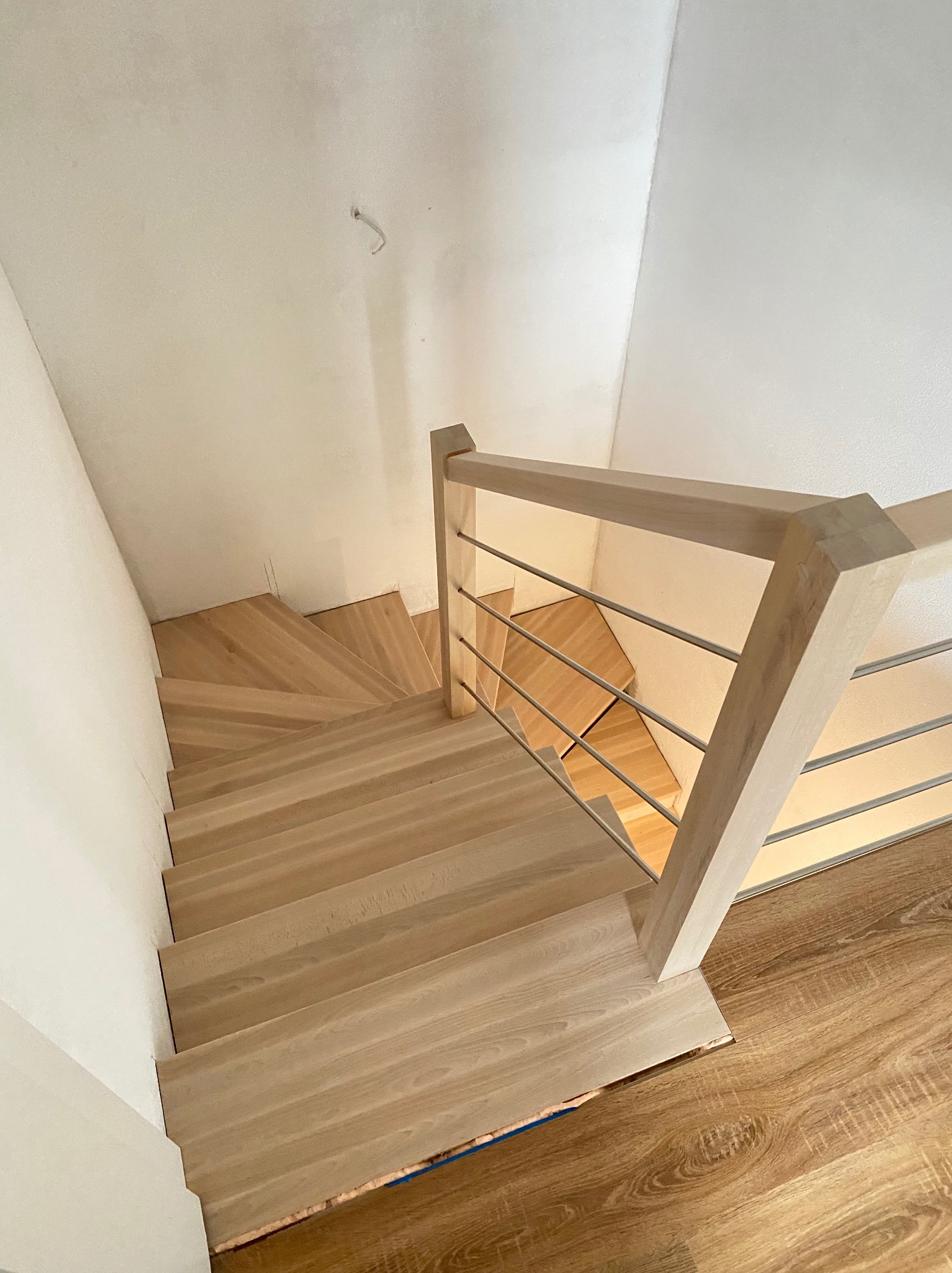 Widok z góry na drewniane schody z jasnego drewna, skręcające w dół, z balustradą z poziomych, metalowych prętów i drewnianych słupków, w pomieszczeniu z białymi ścianami i podłogą imitującą drewno.
