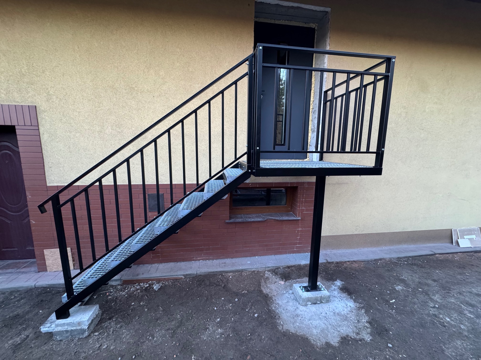Zewnętrzne schody metalowe z platformą, czarna, prosta balustrada, kratownica stopni, betonowe fundamenty, elewacja budynku w tle. Widok z dołu.