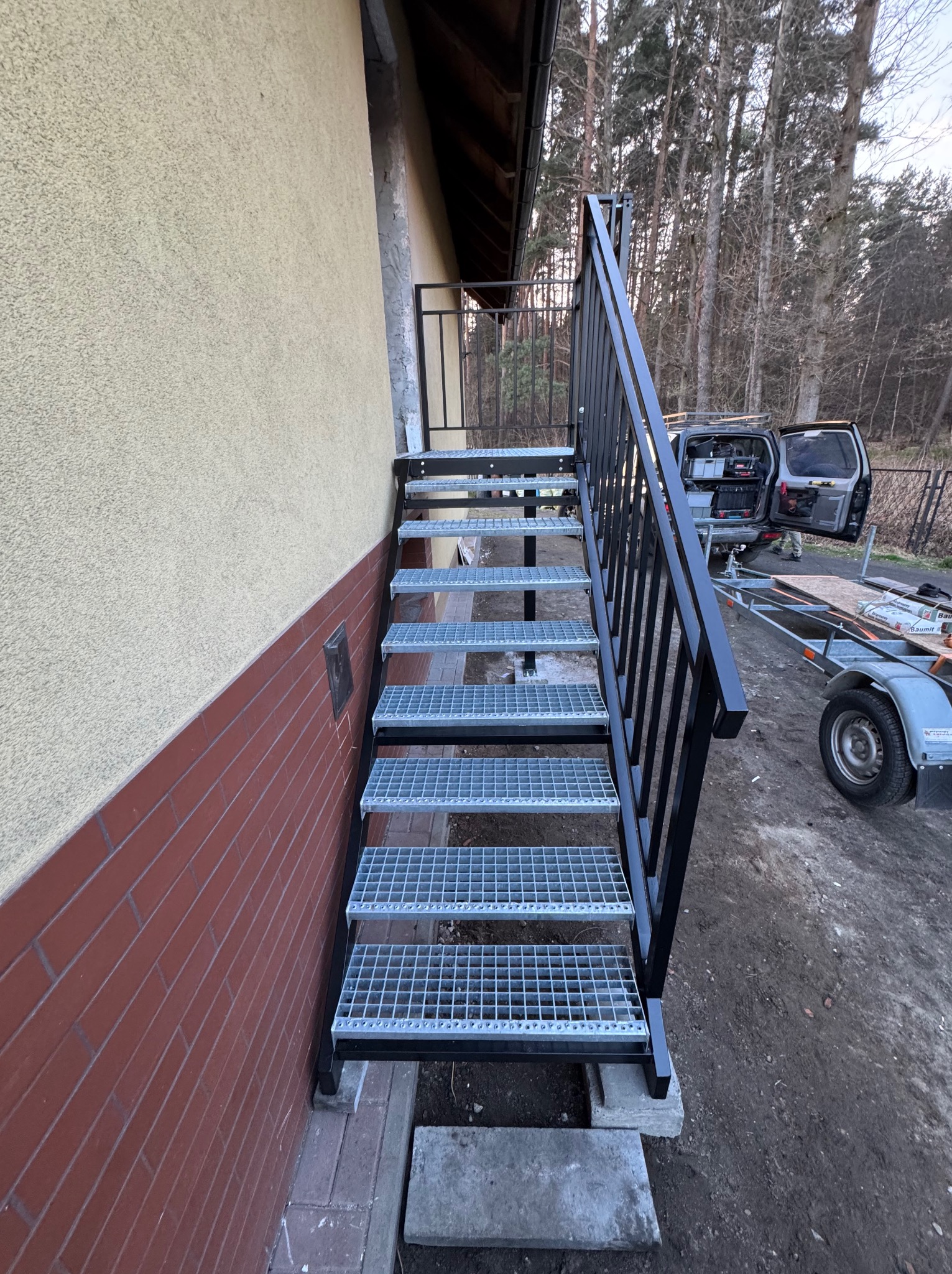 Zewnętrzne schody metalowe z kratowymi stopniami i czarną balustradą, prowadzące do budynku z elewacją w odcieniach beżu i cegły. Widoczny samochód i przyczepka w tle.