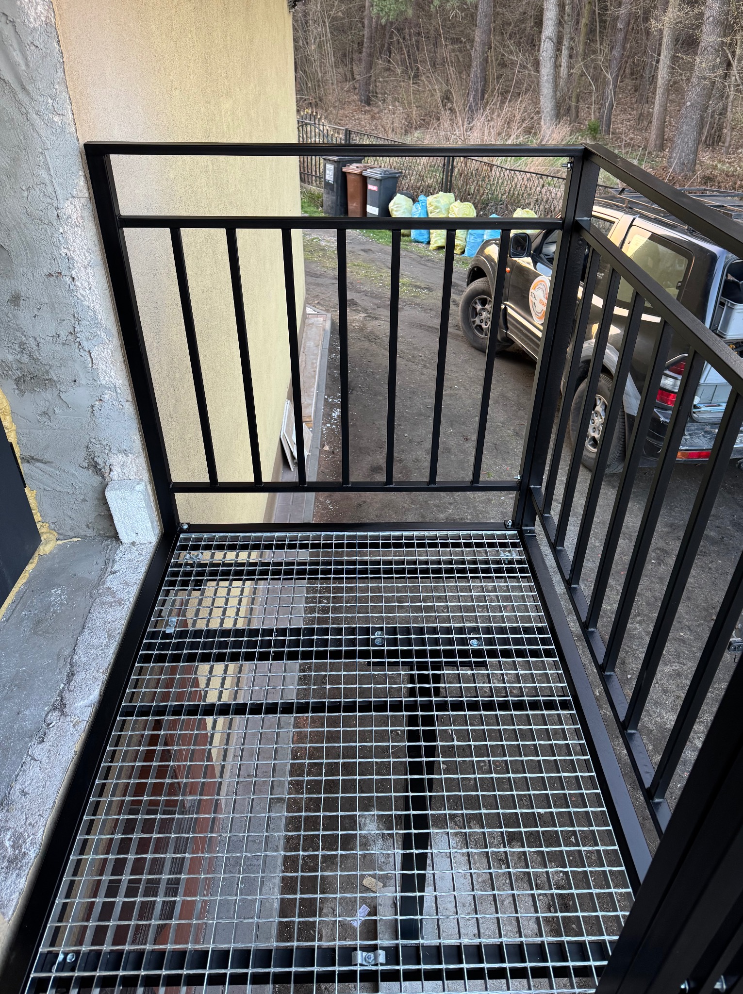 Czarna, metalowa konstrukcja schodów zewnętrznych z kratą, widok z góry. W tle budynek i zaparkowany samochód.