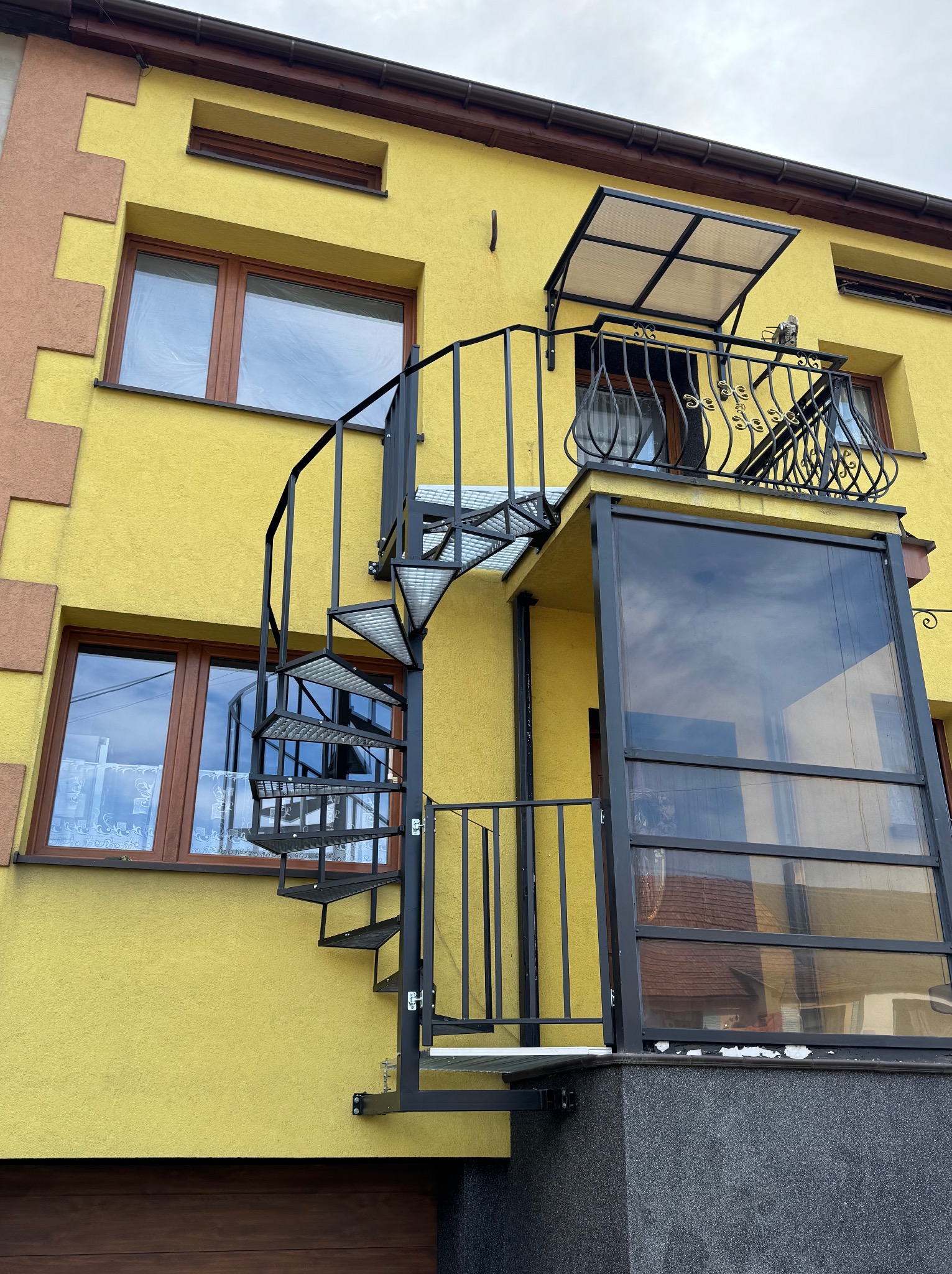 Żółty budynek z zewnętrznymi schodami spiralnymi z metalu, prowadzącymi na balkon z przeszkloną zabudową. Schody z ażurowymi stopniami.