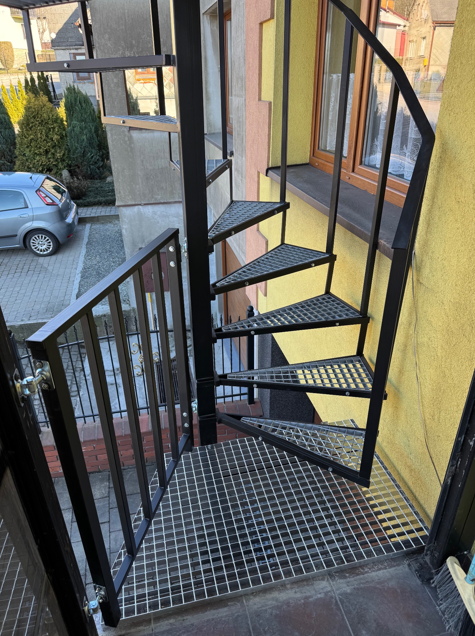 Zewnętrzne schody spiralne z metalu, z ażurowymi stopniami, Bielsko-Biała. Widok z góry na konstrukcję przy budynku z oknem i fragmentem podwórza.