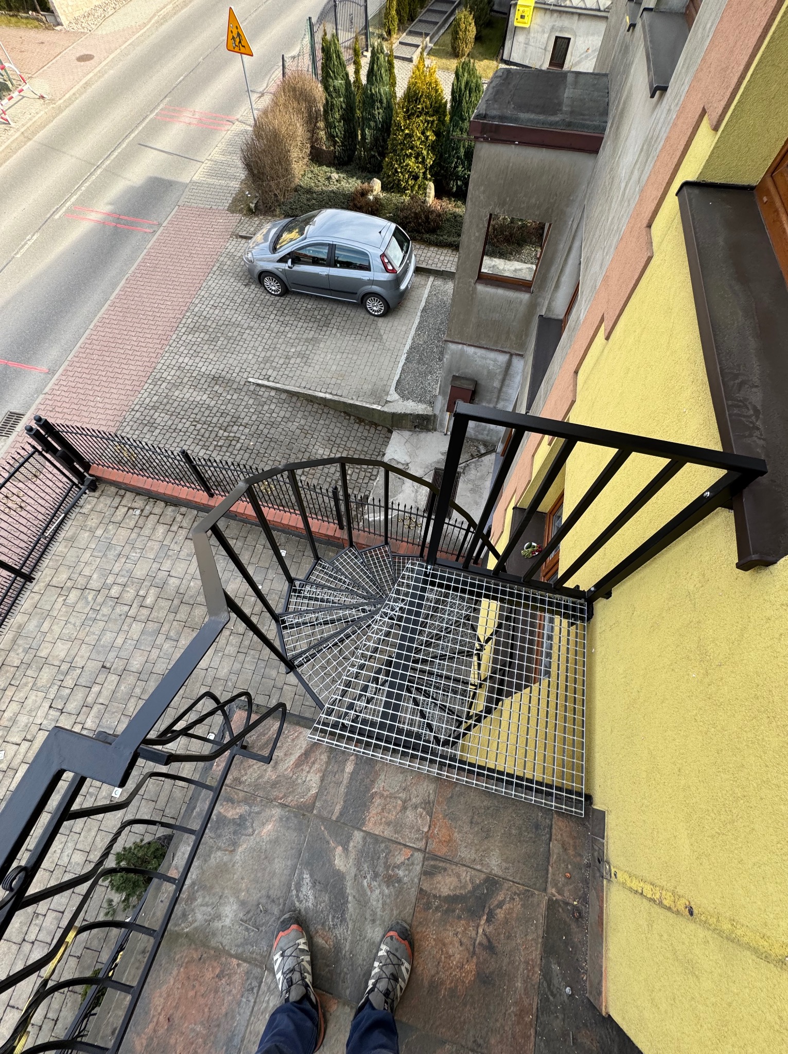 Zewnętrzne schody spiralne z czarną metalową balustradą, widok z góry na tle ulicy z zaparkowanym autem. Nietypowy kadr z perspektywy osoby stojącej na schodach.