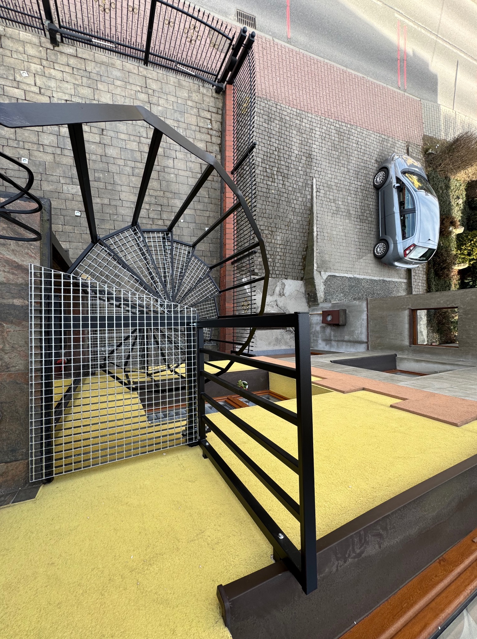 Widok z góry na metalowe schody spiralne z kratą, żółtą nawierzchnią i fragmentem elewacji budynku z zaparkowanym samochodem. Nietypowy kąt ujęcia.