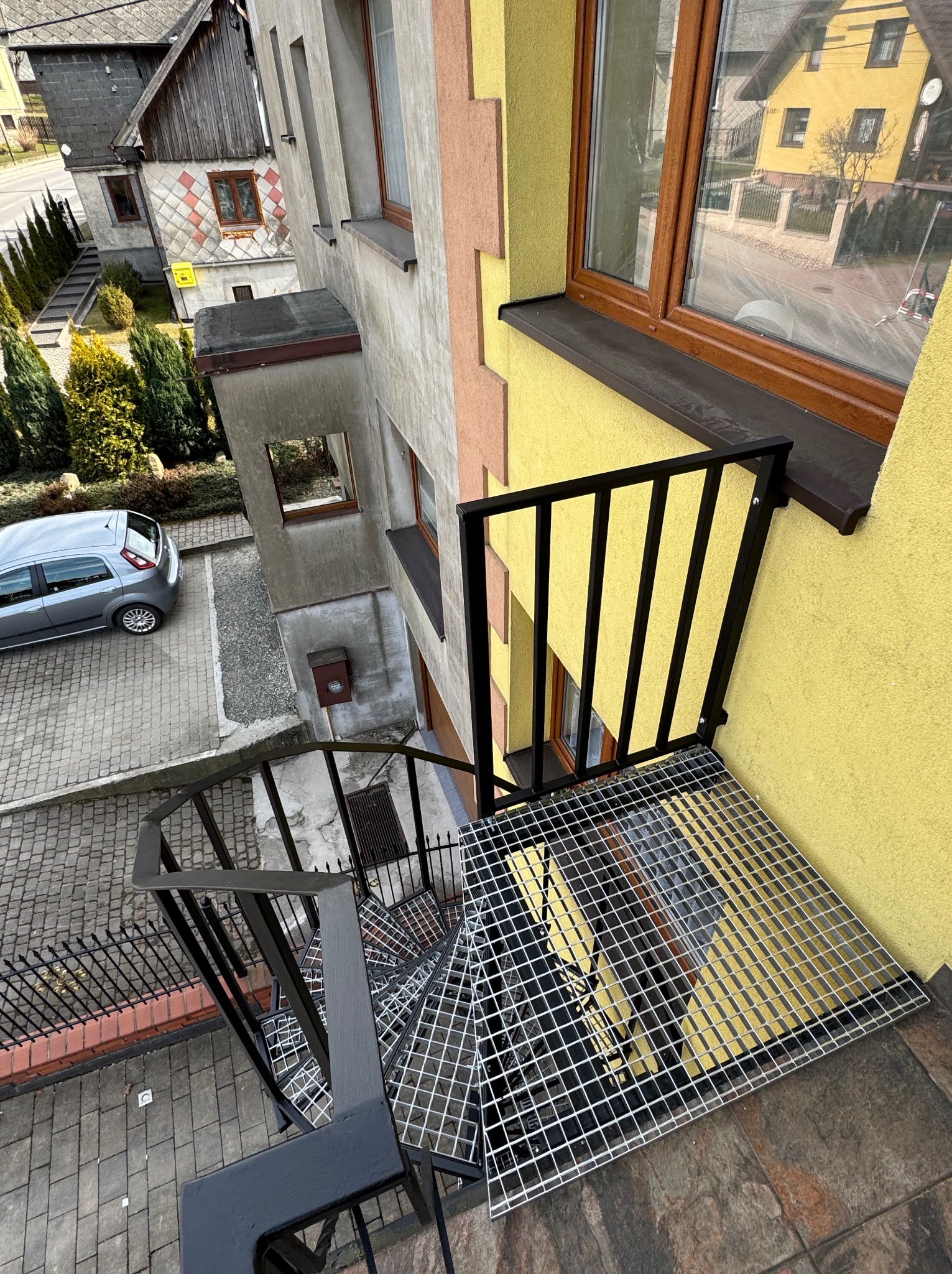 Zewnętrzne schody spiralne z metalową kratką i czarną balustradą, prowadzące z balkonu na dziedziniec. Widok z góry, budynki w tle.