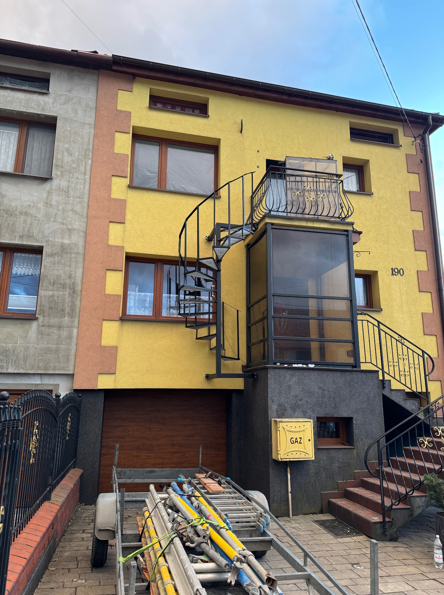 Żółty budynek z metalowymi schodami spiralnymi i przeszkloną werandą na piętrze. Widoczny przyczepka z materiałami budowlanymi i brama wjazdowa.