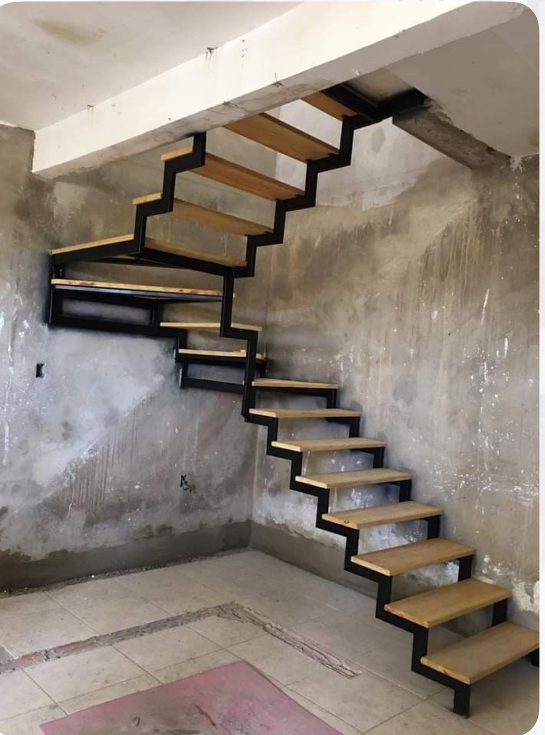 Schody z drewnianymi stopniami i czarną metalową konstrukcją, zamontowane w surowym wnętrzu z betonowymi ścianami. Widok z dołu, ukazujący konstrukcję schodów.
